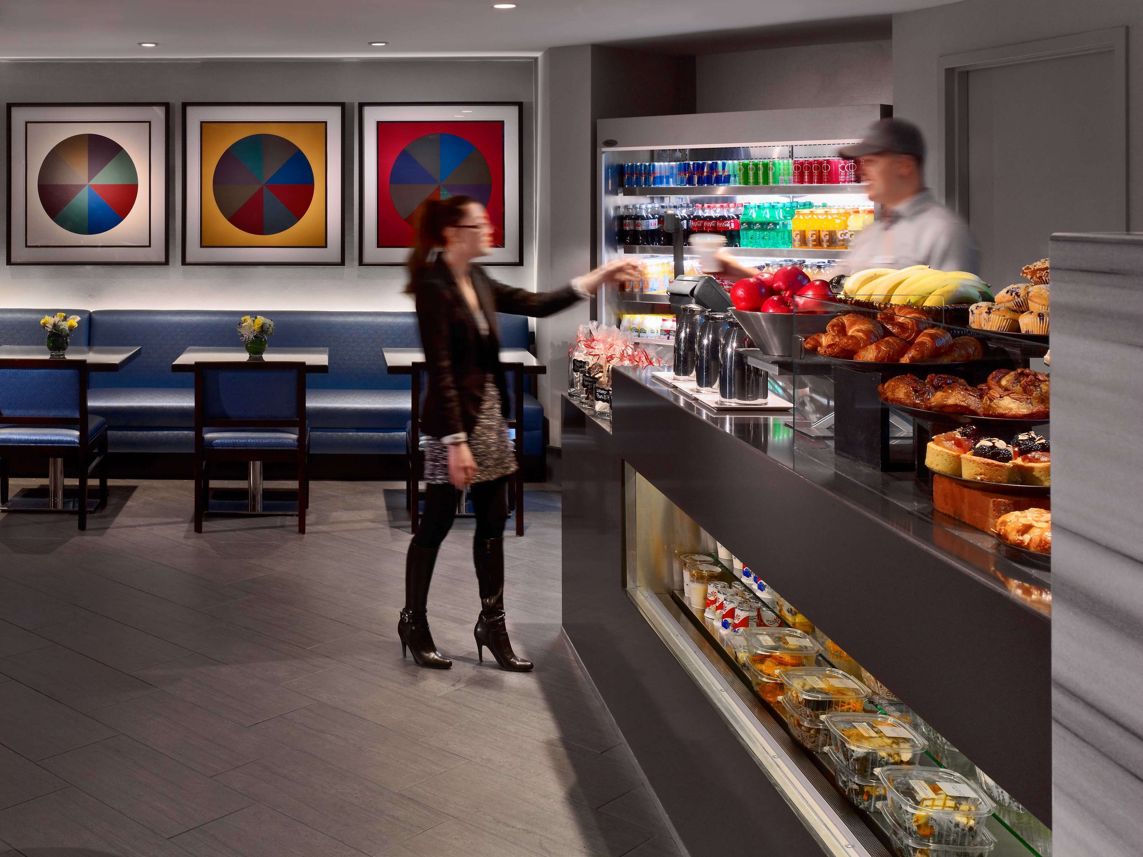 A modern cafe with a counter displaying pastries and beverages, a seating area with blue benches, and framed abstract art on the wall.