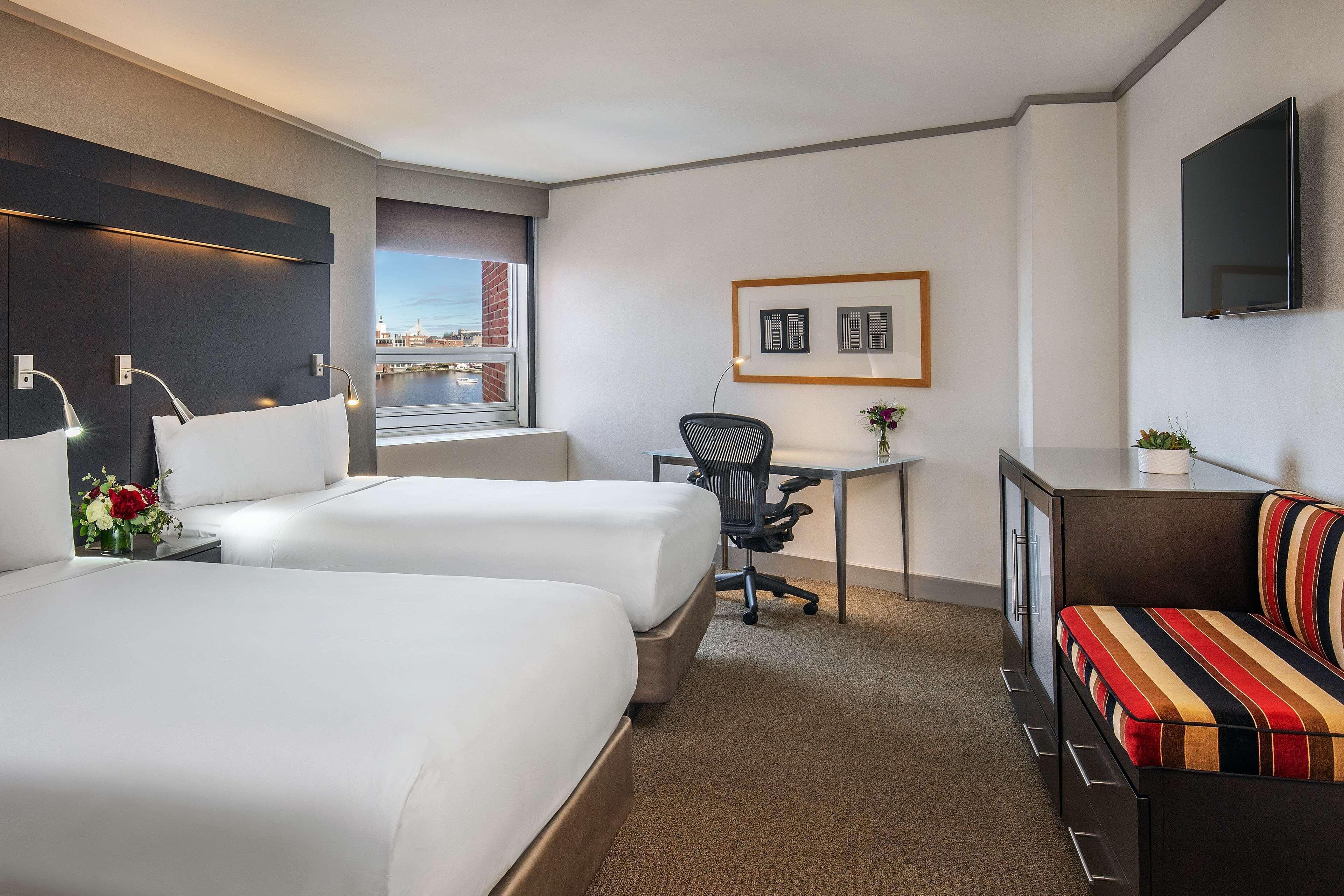 A hotel room with two beds, a desk, a chair, a TV, and a view of a cityscape.