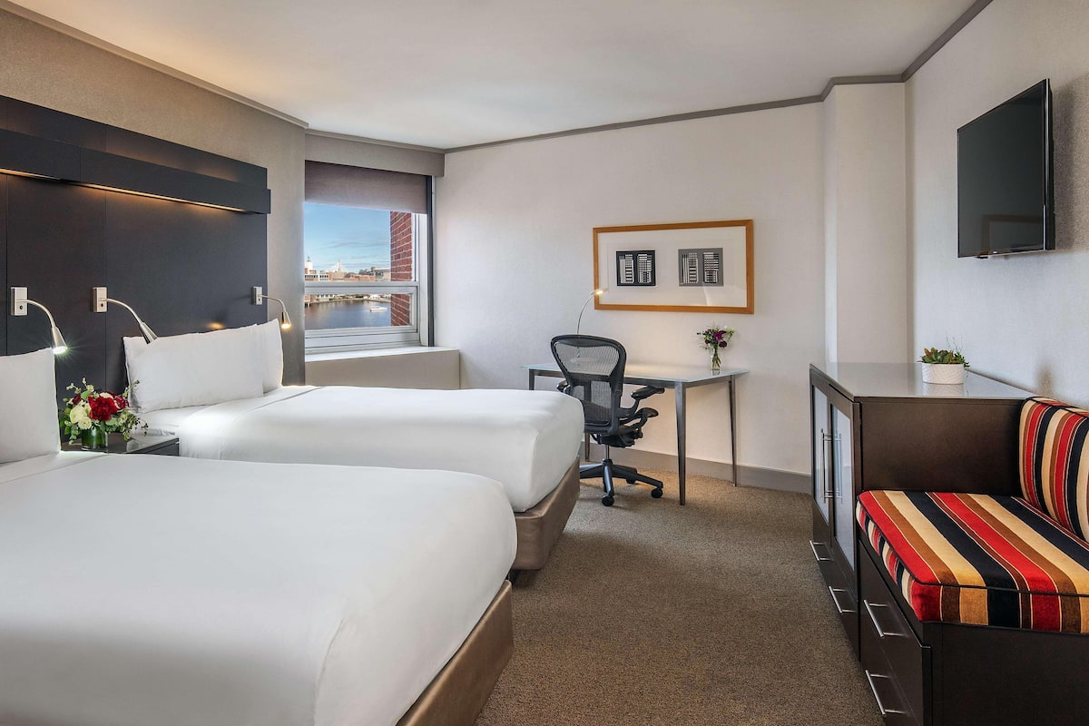 A hotel room with two beds, a desk, a chair, a TV, and a view of a cityscape.