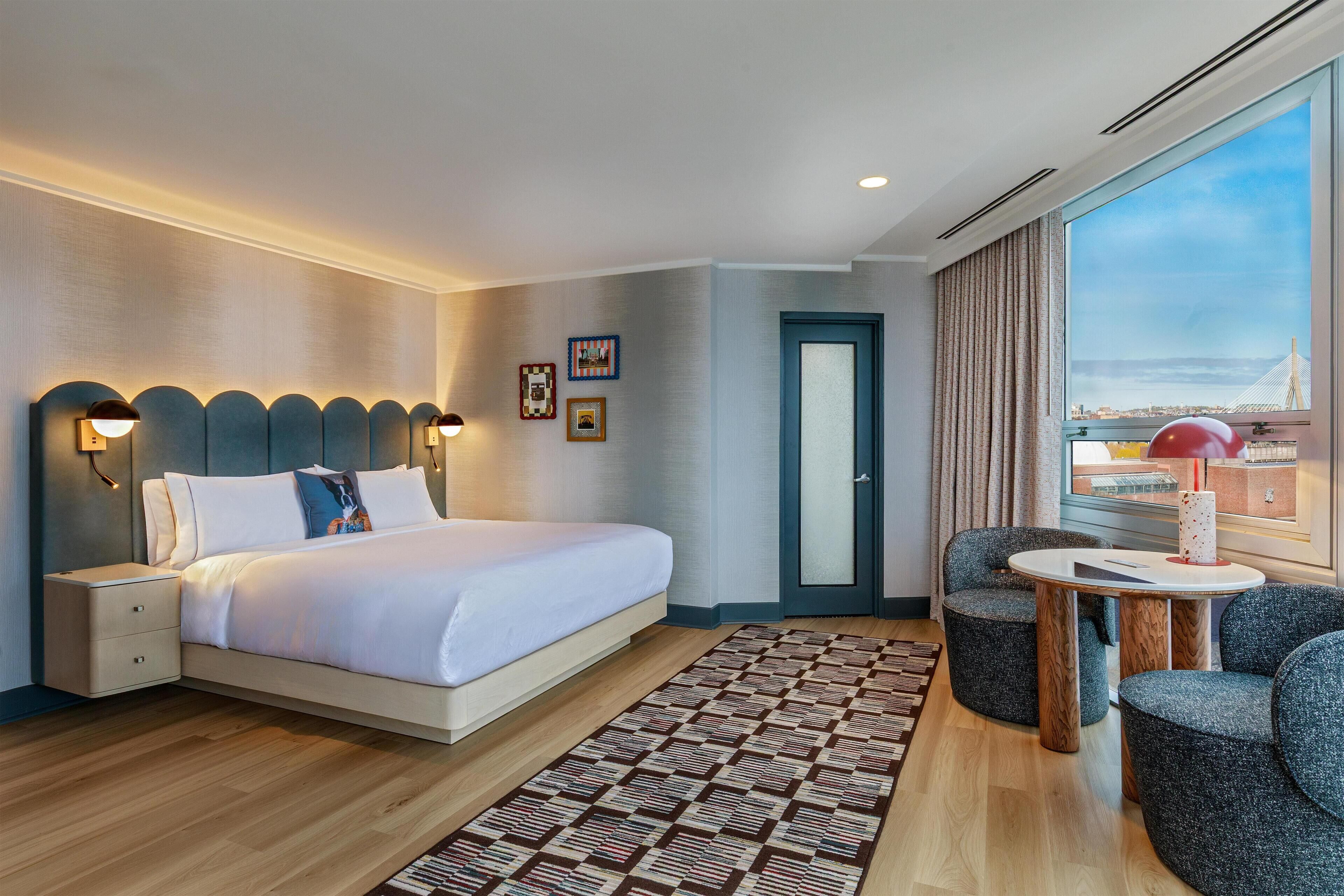A modern hotel room with a large bed, bedside lamps, a small table, and a view of a cityscape.