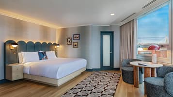 A modern hotel room with a large bed, bedside lamps, a small table, and a view of a cityscape.