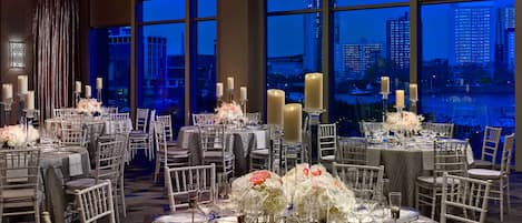 Un salón de banquetes con mesas redondas dispuestas para cenar, grandes ventanales que ofrecen vistas a la ciudad y elegantes centros de mesa con flores.