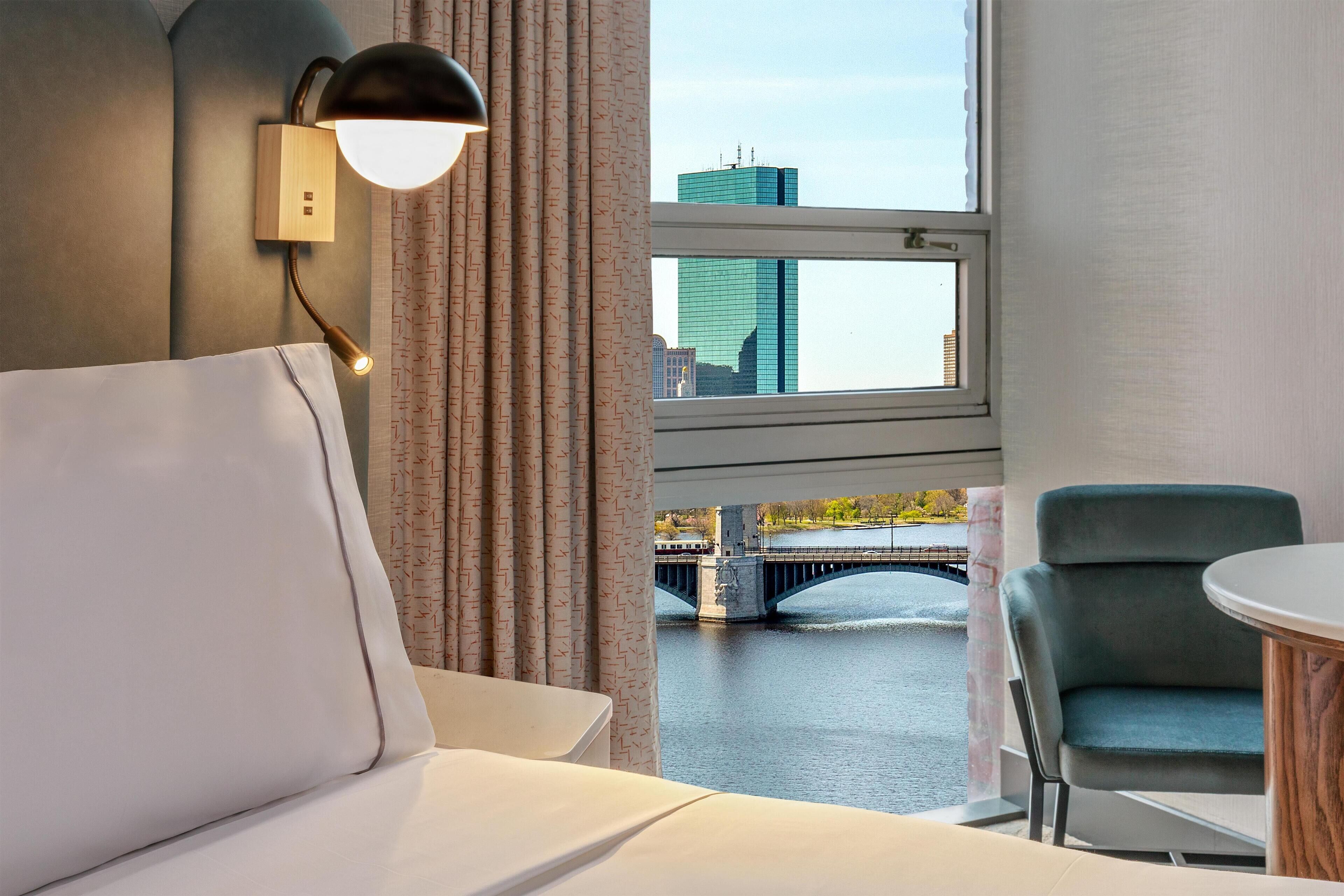 Ein Hotelzimmer mit Bett, Stuhl, Tisch, Lampe und einem Fenster mit Blick auf eine Brücke und Gebäude.