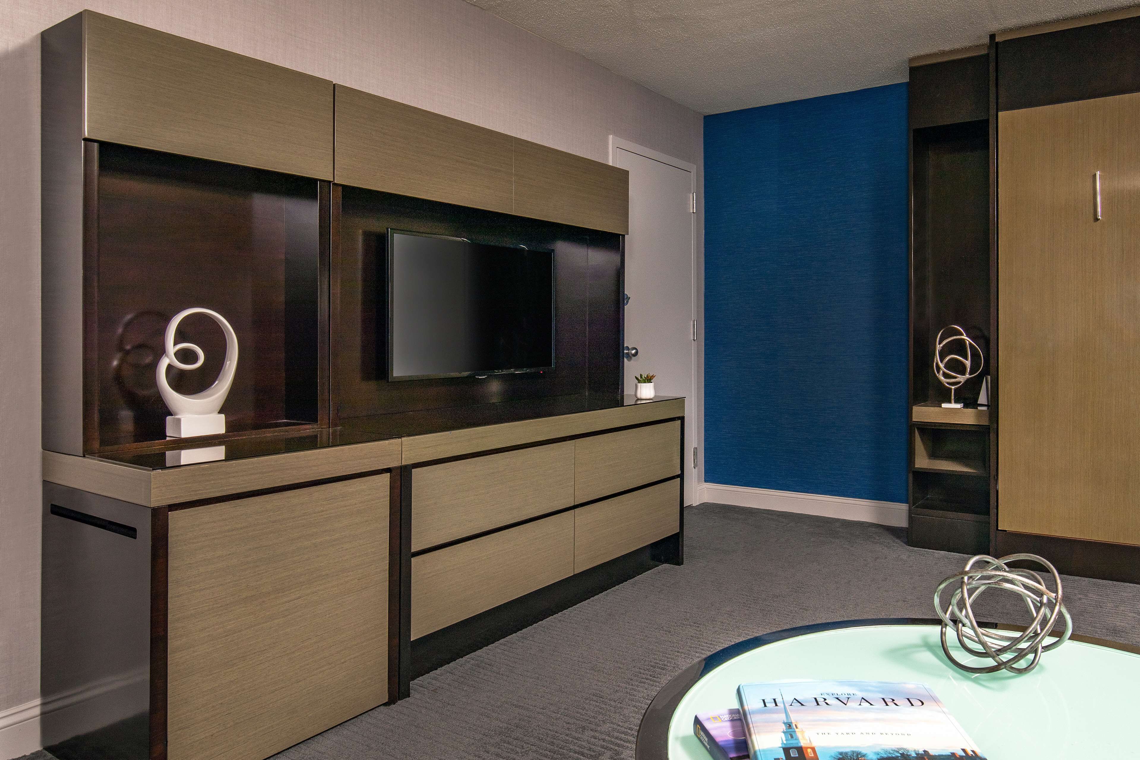 A modern hotel room with a large flat-screen TV, a desk with a magazine, and a blue accent wall.