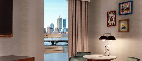 A hotel room with a large window, a desk, two chairs, a round table, and a lamp.