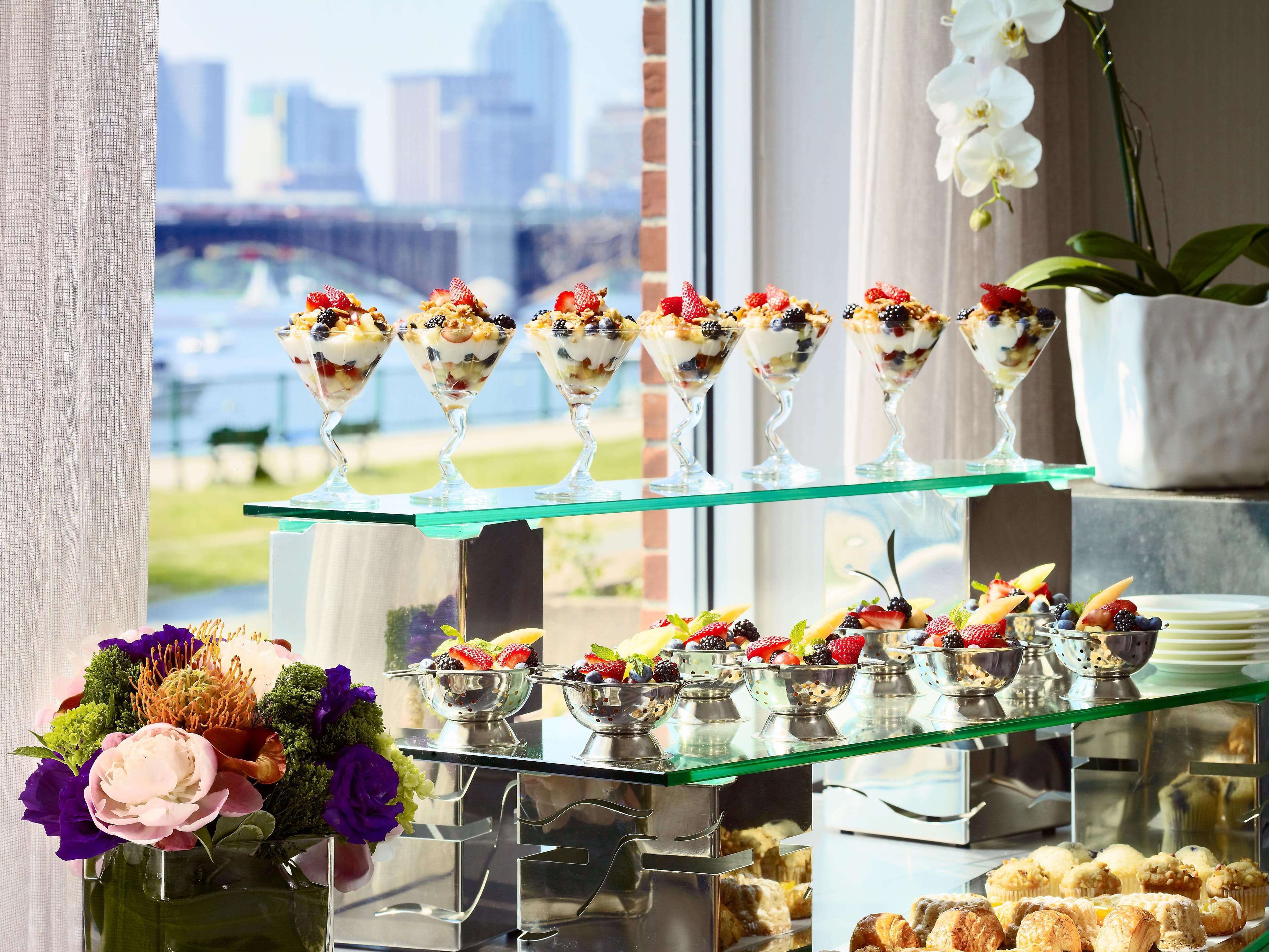 A dessert buffet with various fruit and yogurt parfaits on glass shelves, a floral arrangement, and pastries.