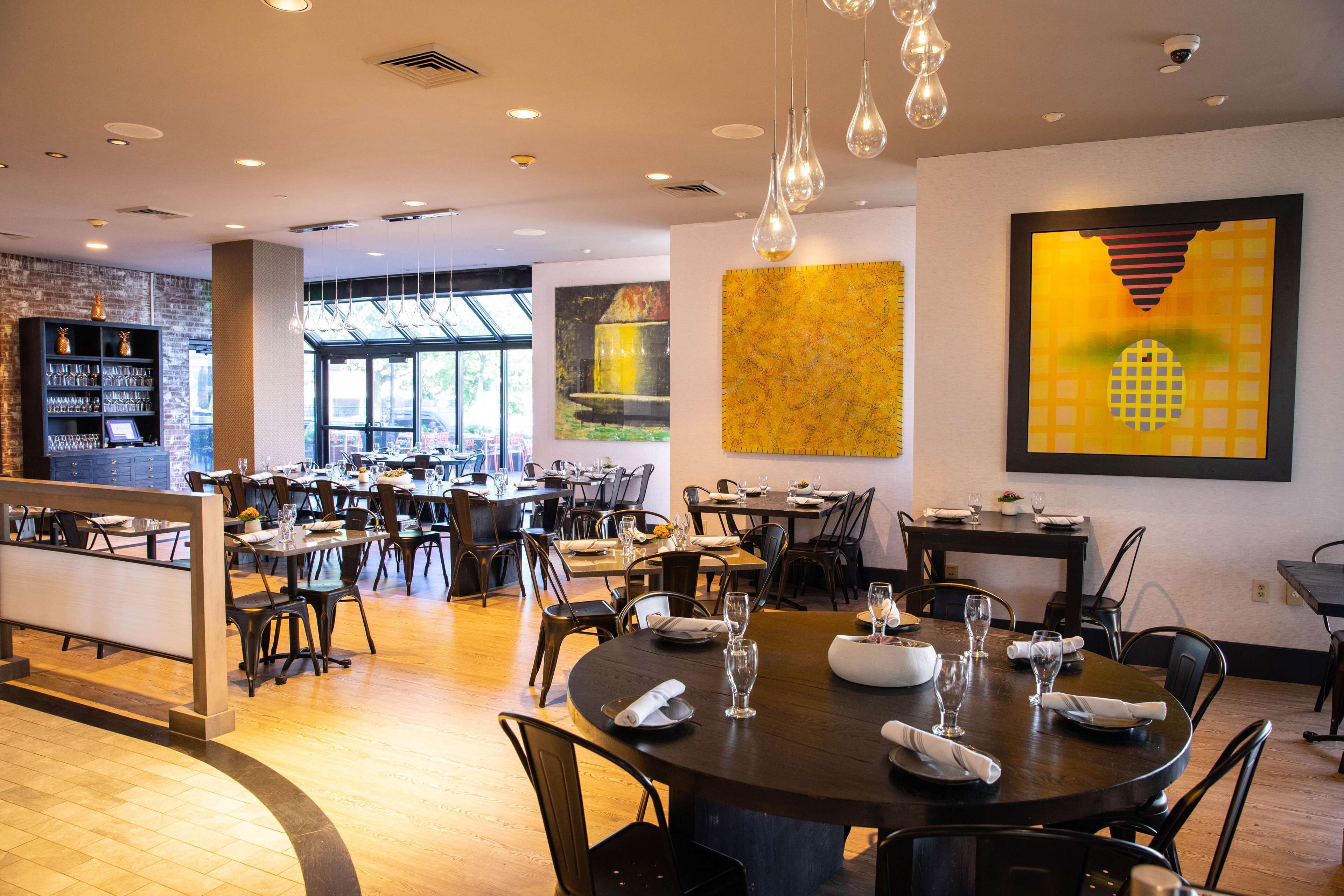 Un comedor moderno con varias mesas y sillas, una zona de bar con estanterías, y ventanas grandes que dejan entrar luz natural.