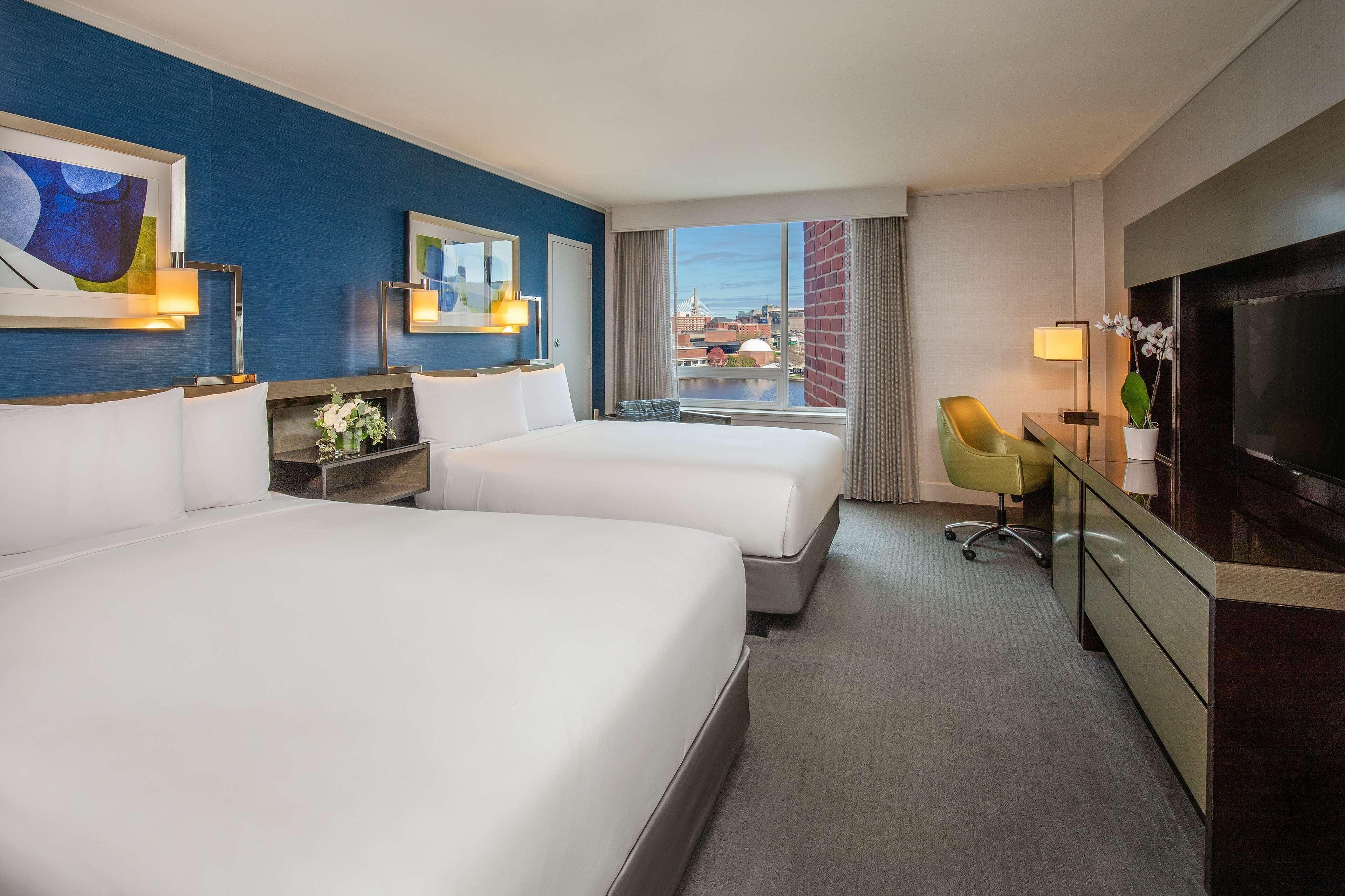 A hotel room with two beds, a desk, a TV, and a view of a cityscape.