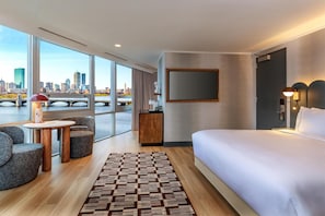 Quarto de hotel moderno com uma cama grande, área de estar com cadeiras, uma pequena mesa, televisão e vista para o horizonte urbano através de grandes janelas.