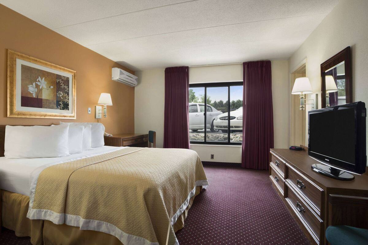 Habitación de hotel con cama, televisor, cómoda, ventana con cortinas y un cuadro en la pared.
