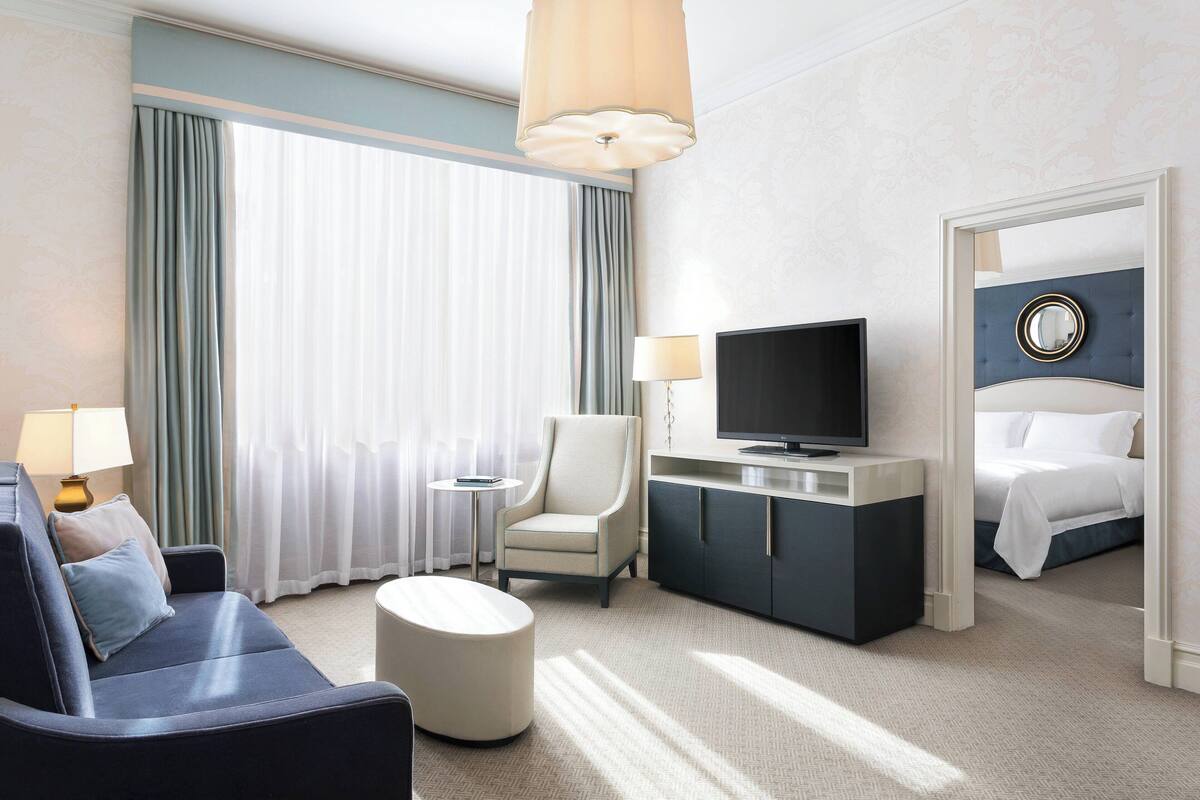 A hotel room with a sofa, armchair, ottoman, TV, and a bed.