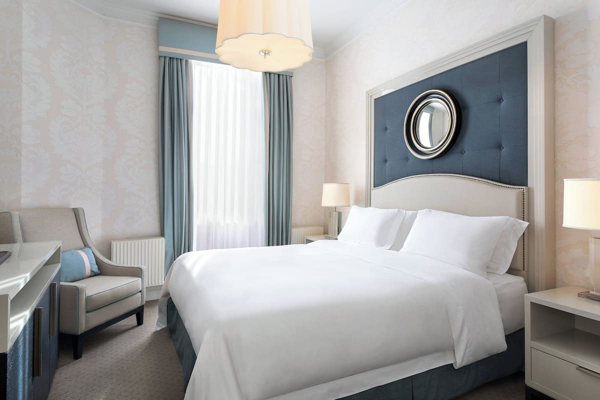 A hotel room with a large bed, a chair, a desk, and a nightstand with a lamp.