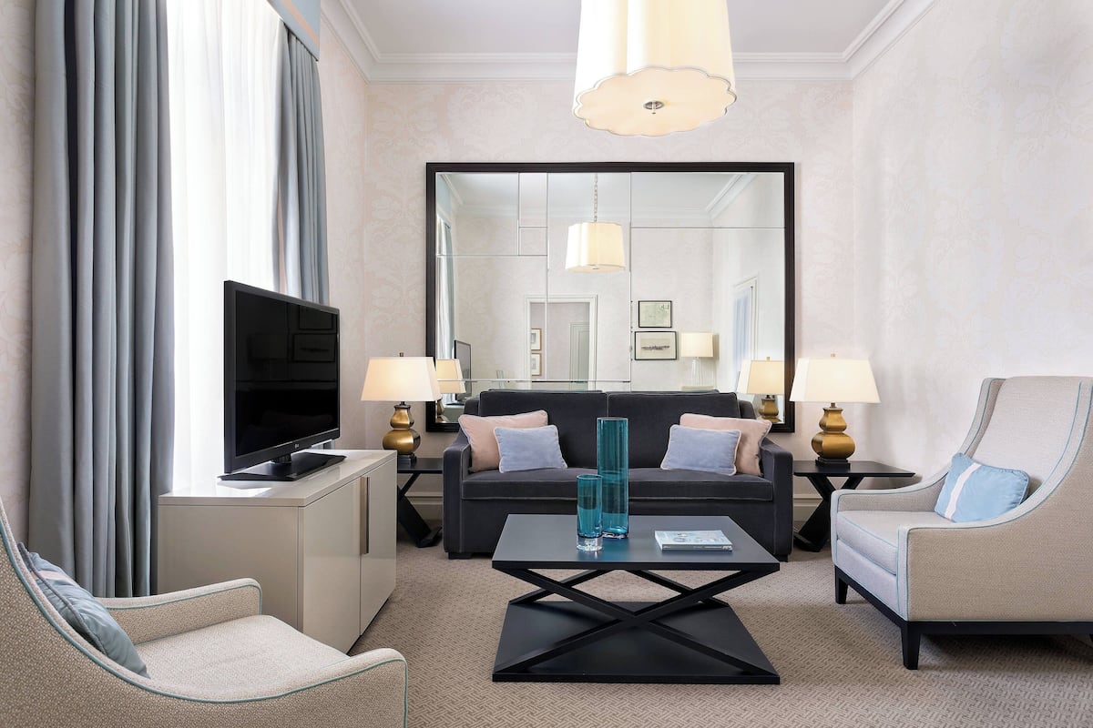 A modern hotel room with a flat-screen TV, a sofa, two armchairs, a coffee table, and a large mirror.