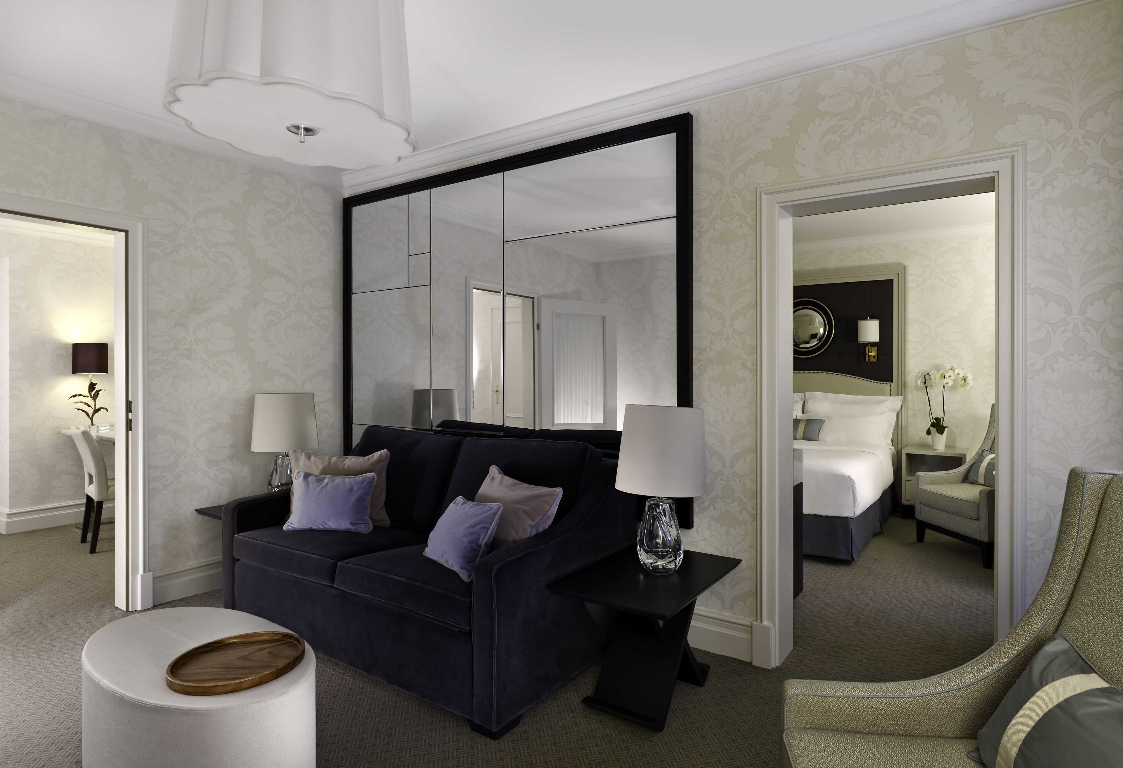 Ein modernes Hotelzimmer mit einem dunkelblauen Sofa, einem runden Hocker, einem großen Spiegel und einem separaten Schlafbereich.