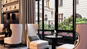 Una moderna hall d'albergo con due poltrone bianche, un tavolino rotondo e un'ampia finestra che si affaccia sul giardino e sulla facciata dell'edificio.
