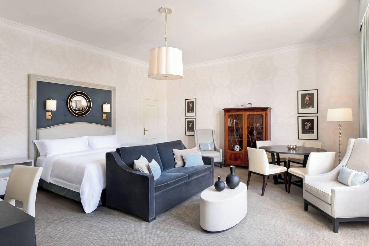 A hotel room with a bed, a dark sofa, a white armchair, a round coffee table, and a wooden cabinet.