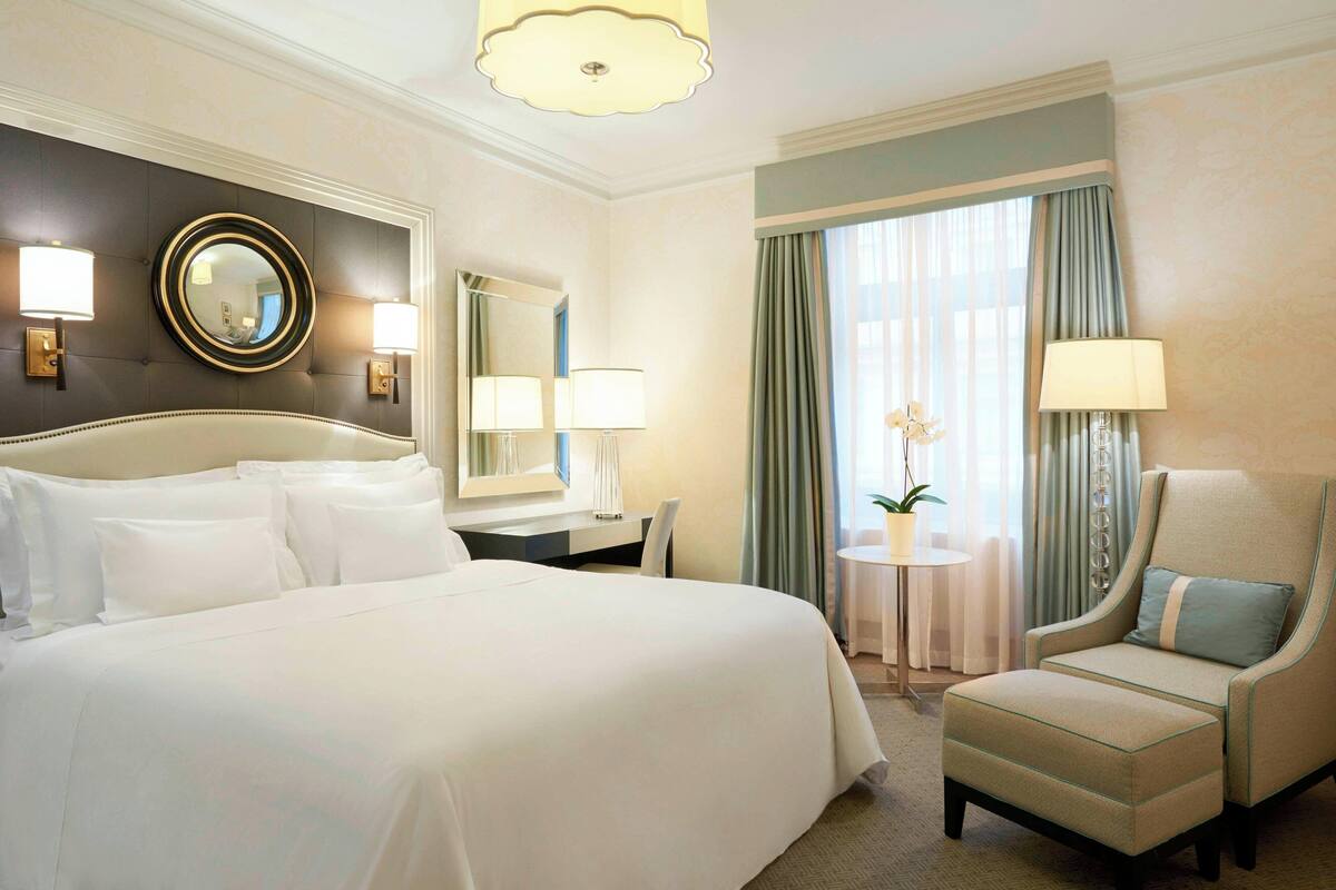 A hotel room with a large bed, a chair, a desk, a round mirror, and a small table with a lamp.