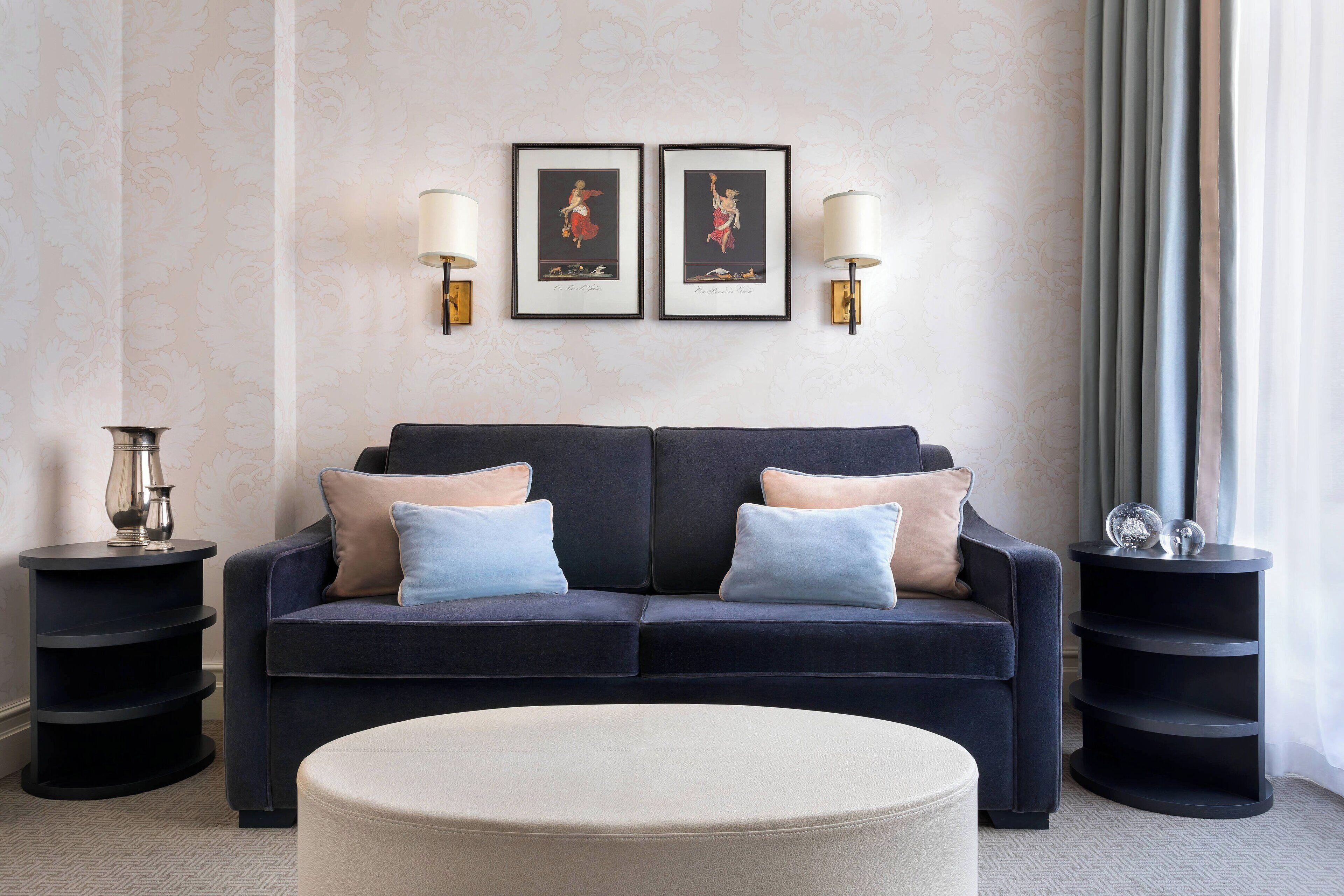 A modern living room with a dark blue sofa, two side tables, a round ottoman, and framed artwork on the wall.