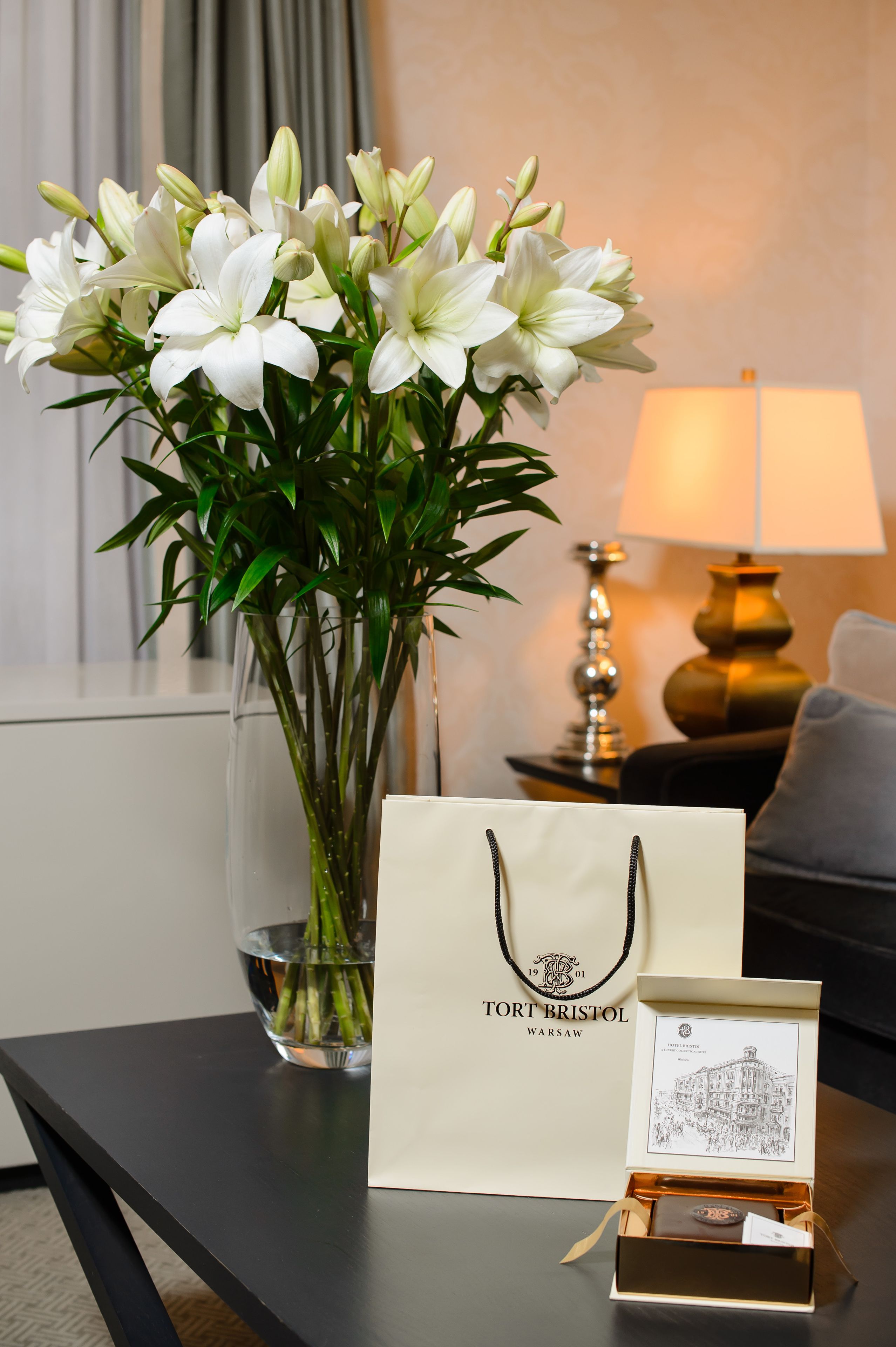 A table with a vase of white lilies, a Torto Bristol Warsaw gift bag, and a small box.
