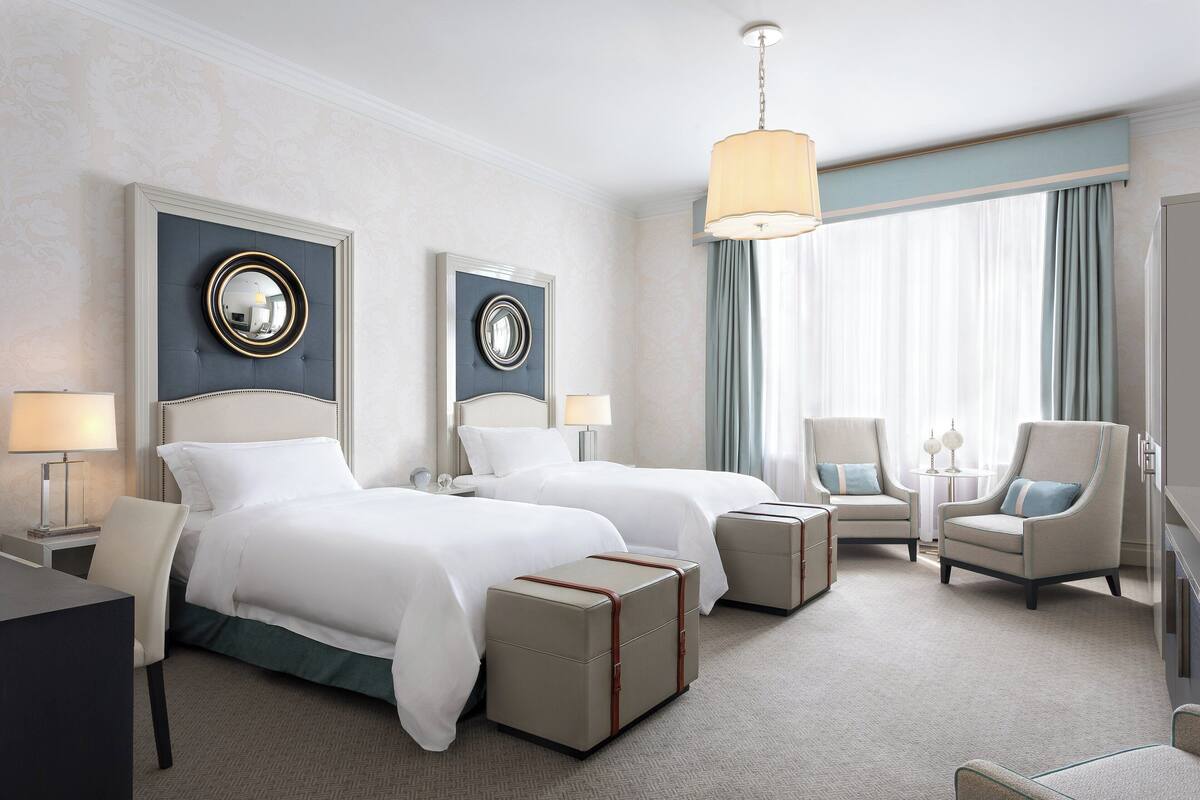 A hotel room with two beds, a desk, chairs, and a large window with curtains.