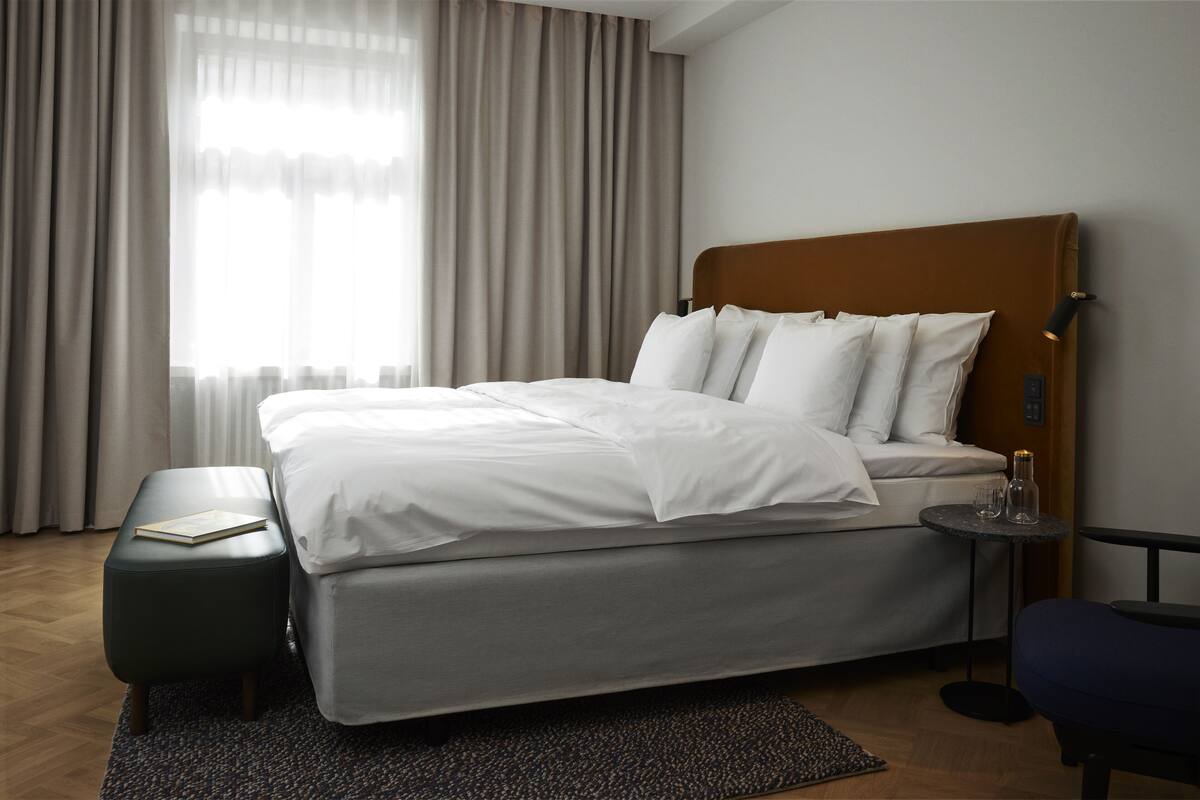 Ein modernes Hotelzimmer mit einem großen Bett, einem kleinen Beistelltisch, einer Bank und Blick auf die Stadt durch transparente Vorhänge.
