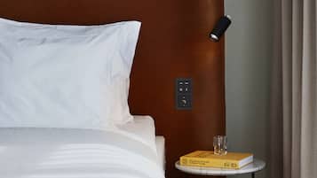 Une chambre d’hôtel avec une tête de lit marron, un lit blanc, une table de chevet avec un livre et un verre, et un panneau de commande au mur.