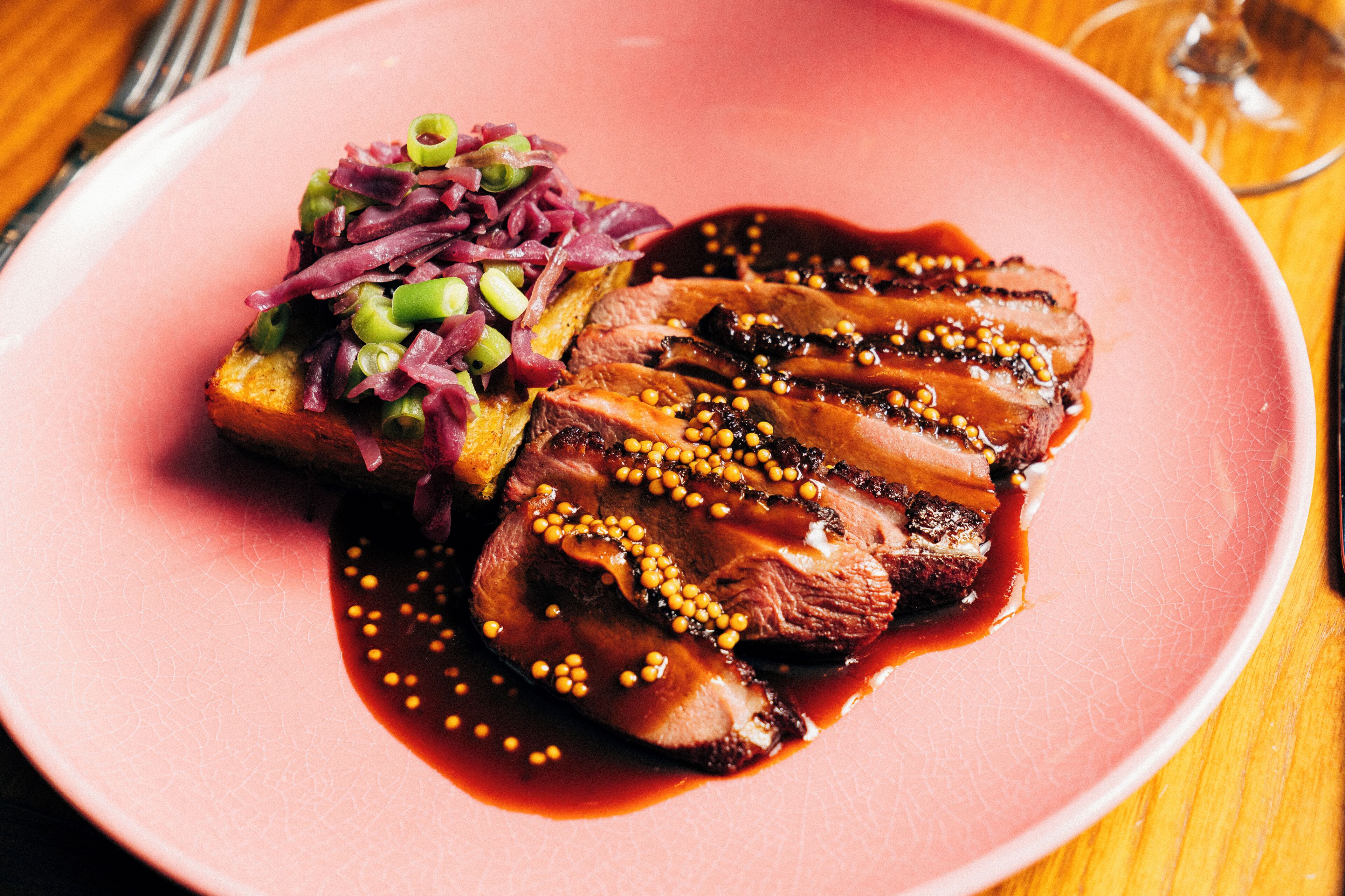 A pink plate with sliced meat, a side of red cabbage, and a piece of toast.