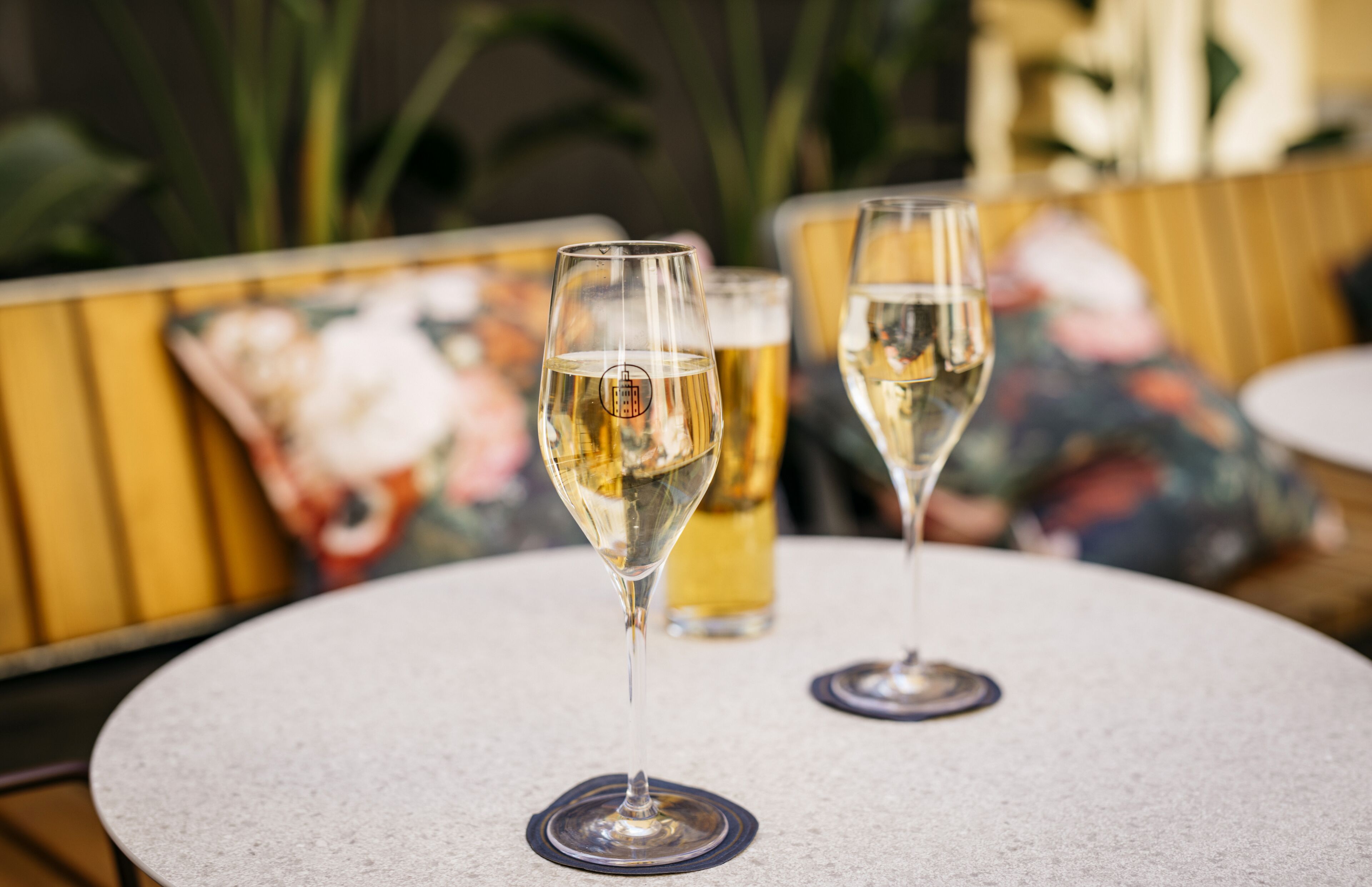 Two glasses of white wine on a round table with a floral cushion in the background.
