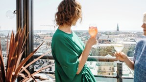 Una persona con un vestido verde sosteniendo una copa de vino, de pie junto a una ventana con vista a la ciudad.