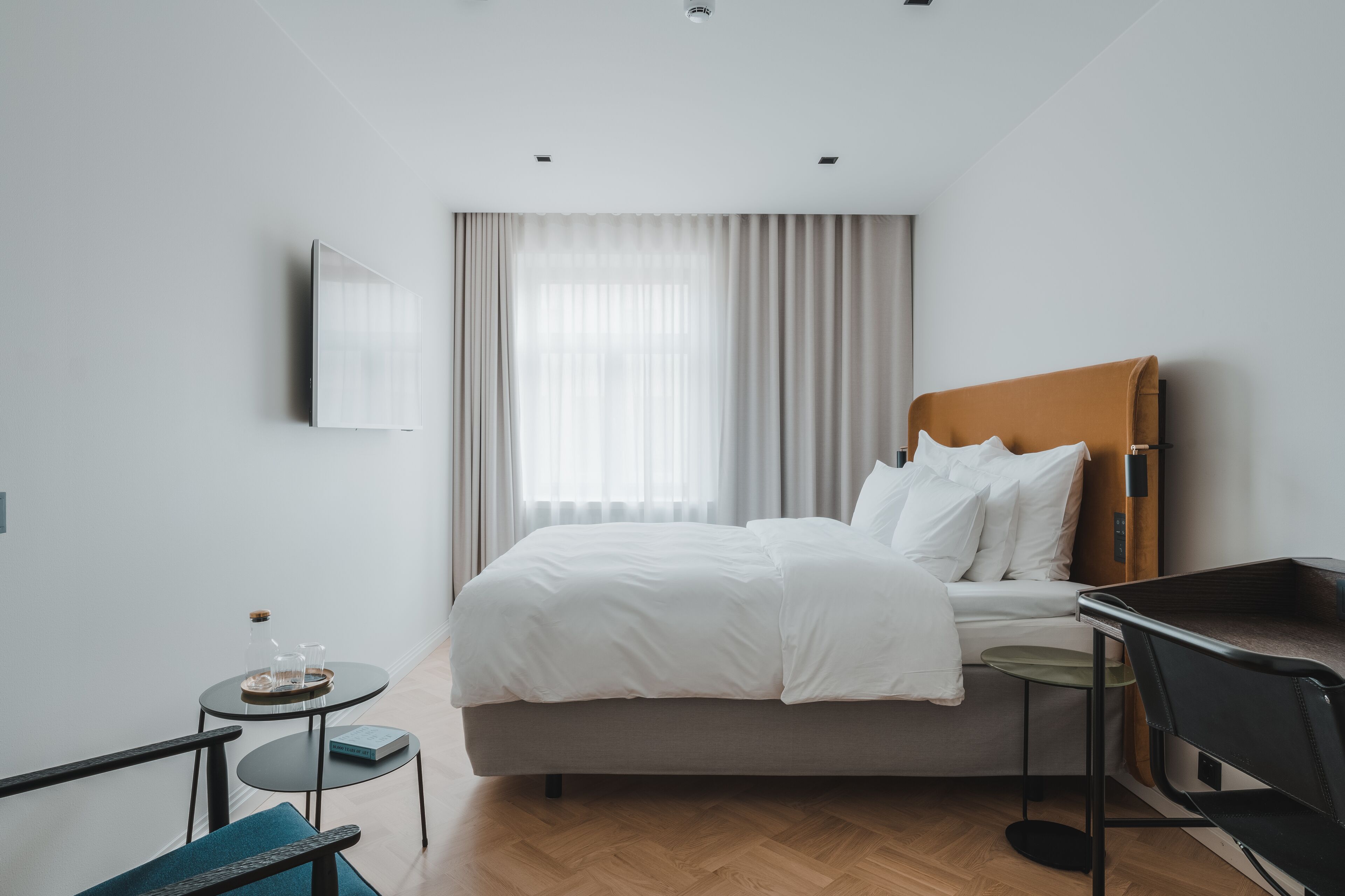 A modern hotel room with a single bed, a small table with a bottle and glasses, a chair, and a desk.
