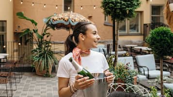 Une femme verse du champagne dans un seau en métal, sur une terrasse extérieure agrémentée de plantes en pot, de meubles de jardin et de guirlandes lumineuses.
