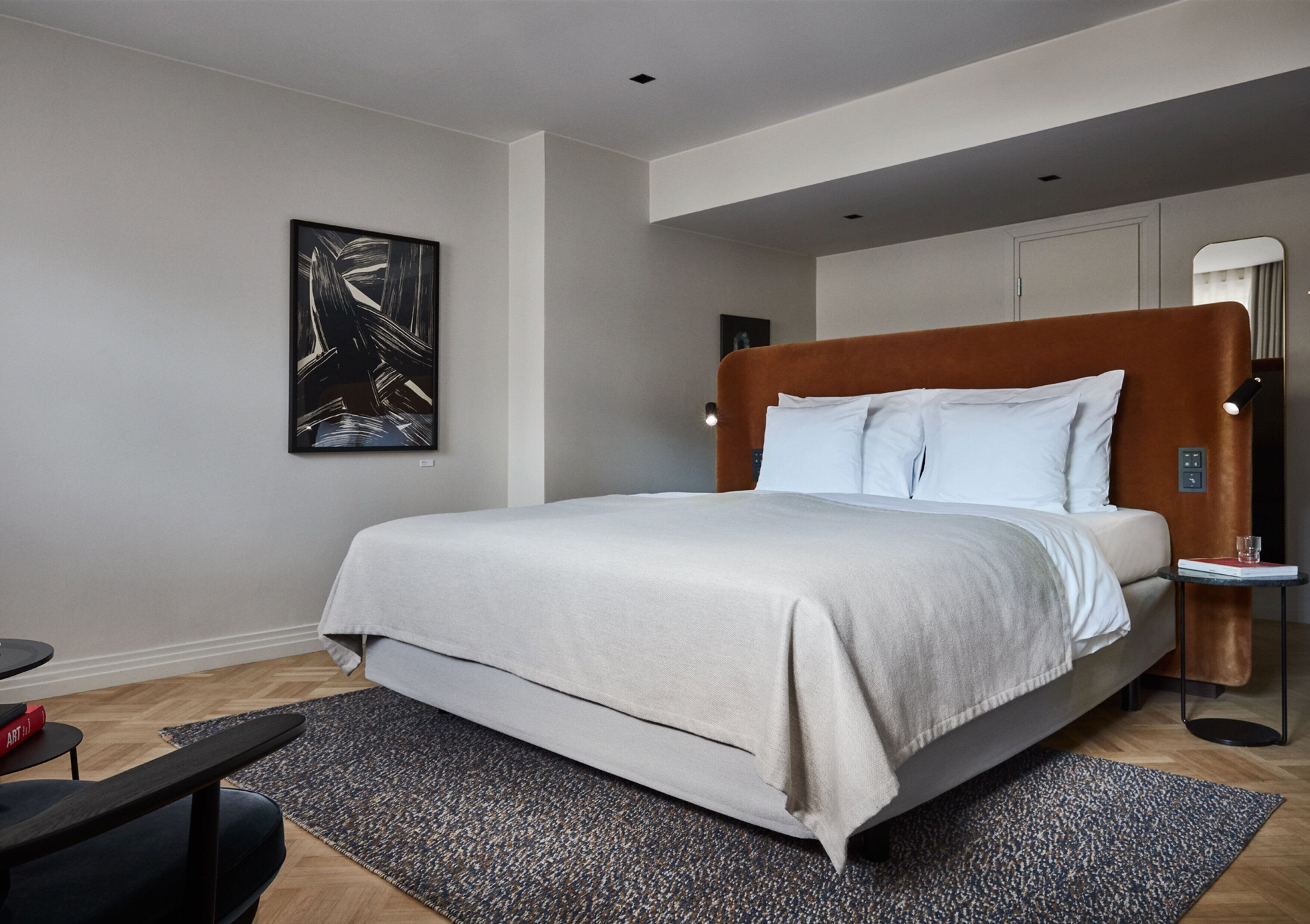 A modern bedroom with a large bed, a side table, a floor lamp, and a painting on the wall.