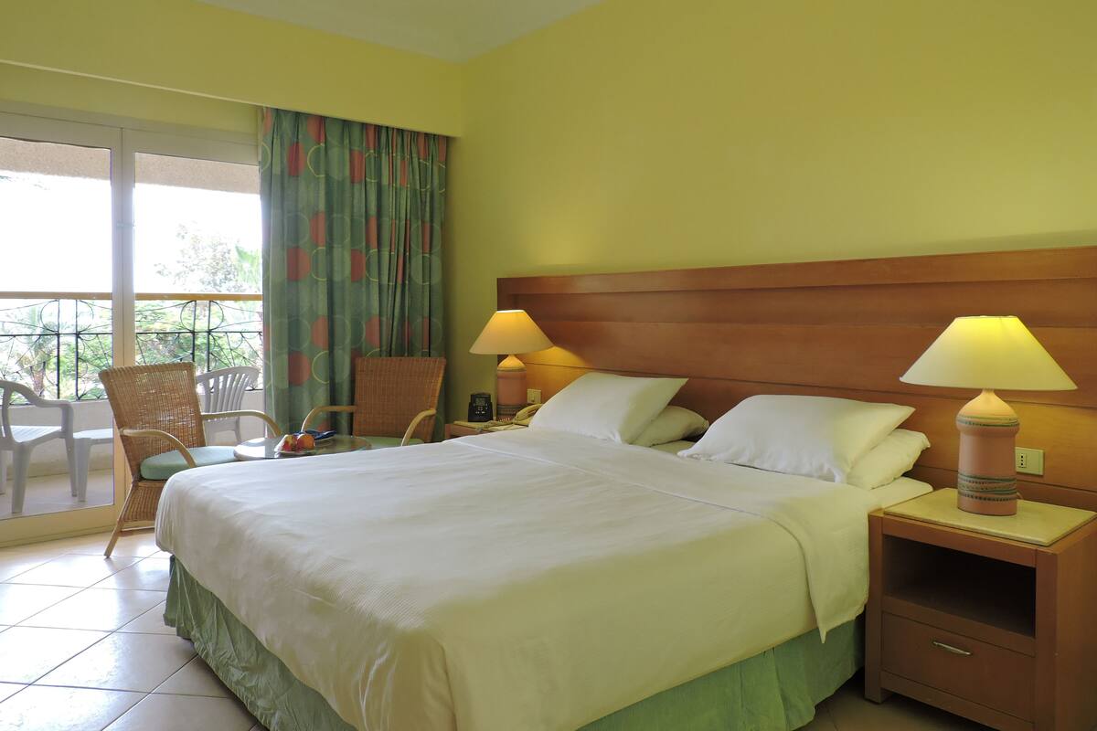 A hotel room with a bed, two bedside tables with lamps, a chair, a balcony with a view, and a curtain.