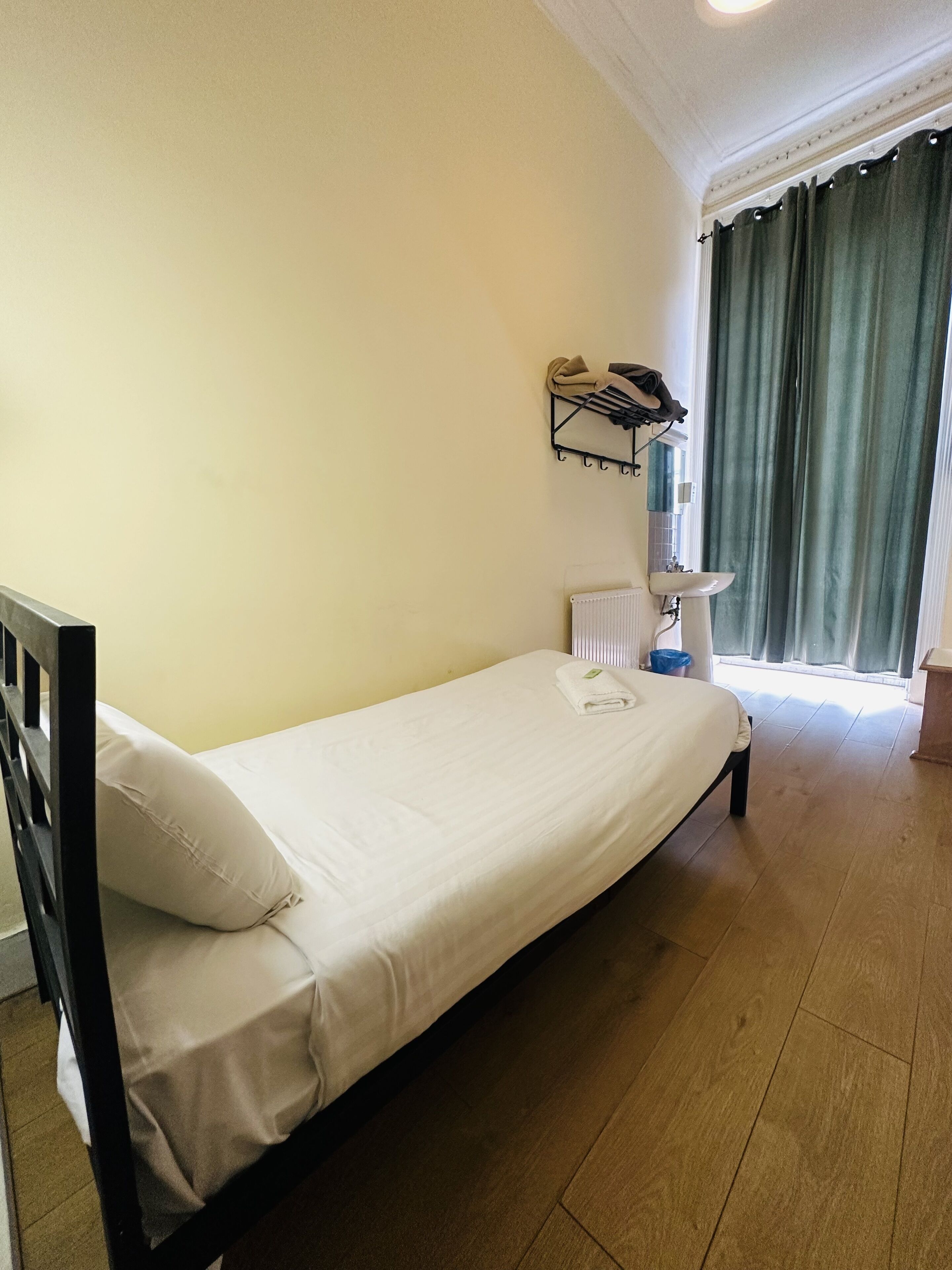 A single bed with a white mattress and a beige pillow, a small bedside table with a lamp, a radiator, and a window with curtains.