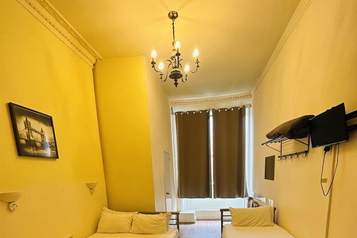 A room with two single beds, a chandelier, a flat-screen TV mounted on the wall, and a window with curtains.