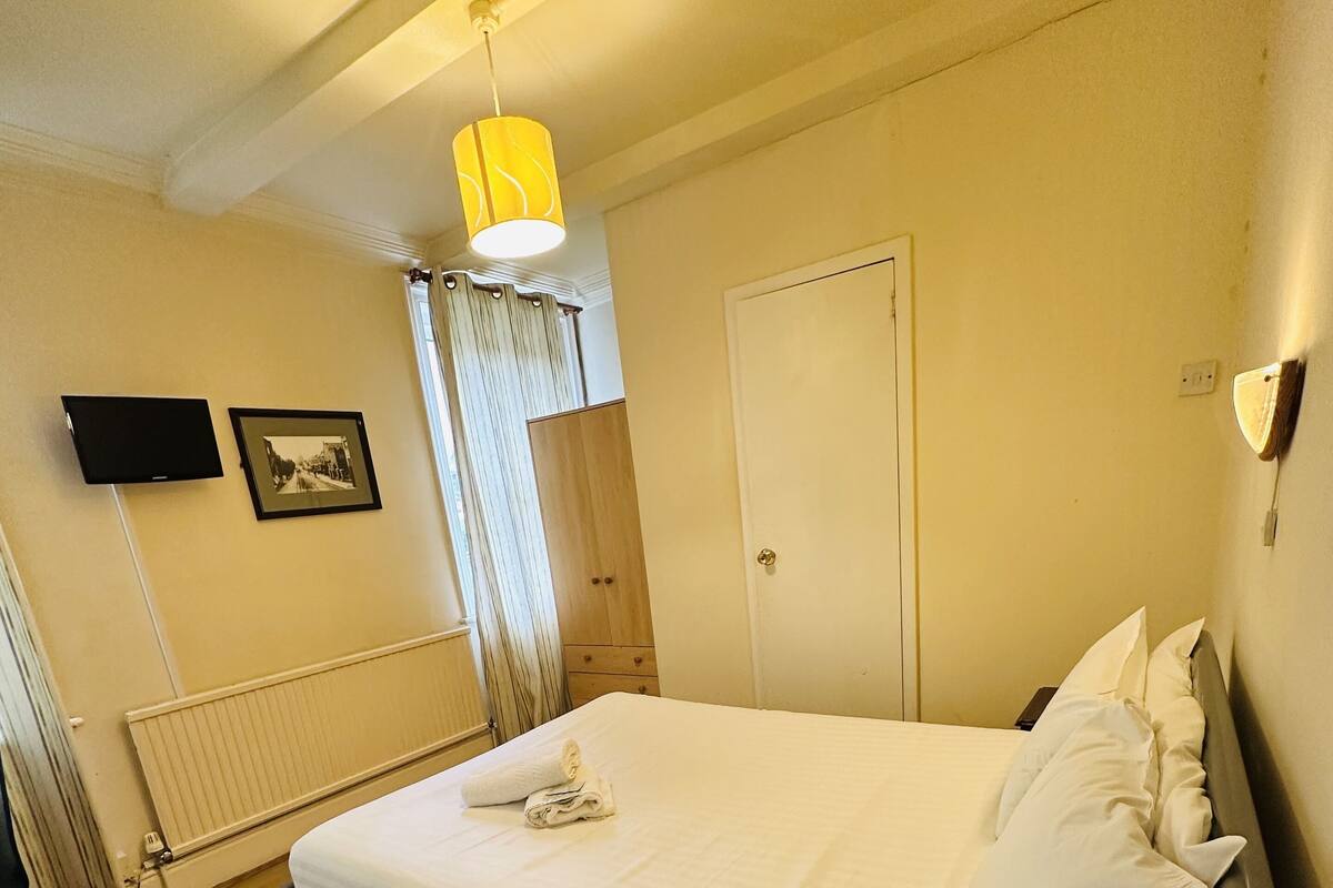 A hotel room with a bed, a flat-screen TV, a radiator, and a window with curtains.