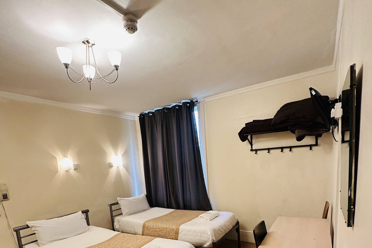 A room with two beds, a desk, and a chair. There is a ceiling fan, wall-mounted lights, and a coat rack.