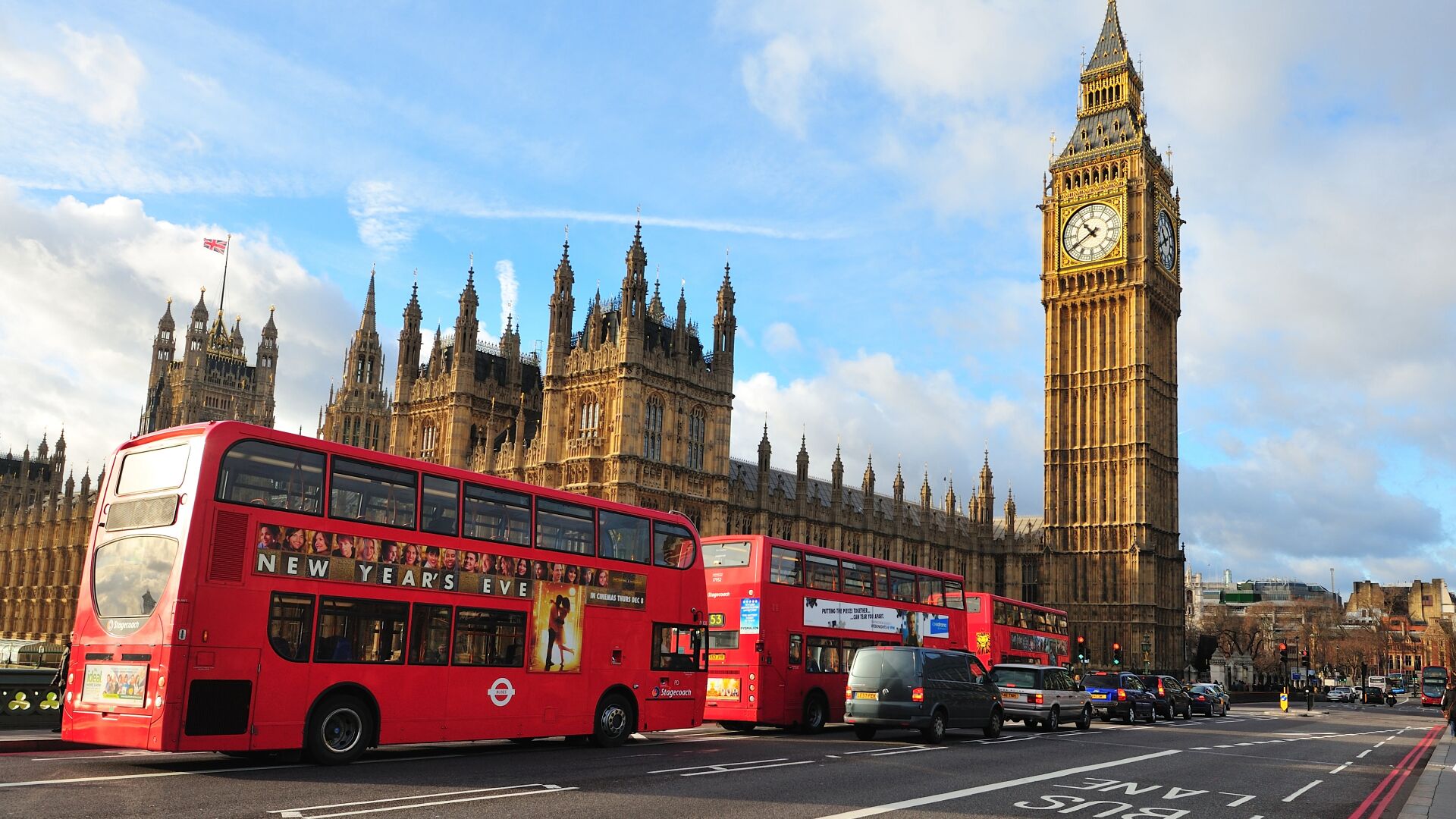 A red double-decker bus with advertisements, a clock tower, and a historic building in the background.