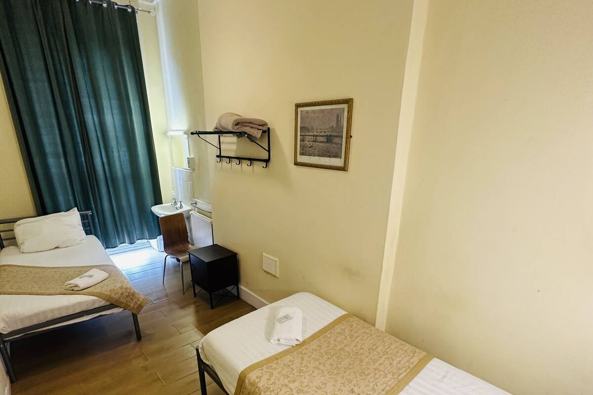 A hotel room with two single beds, a desk, a chair, a small table, a radiator, and a picture on the wall.