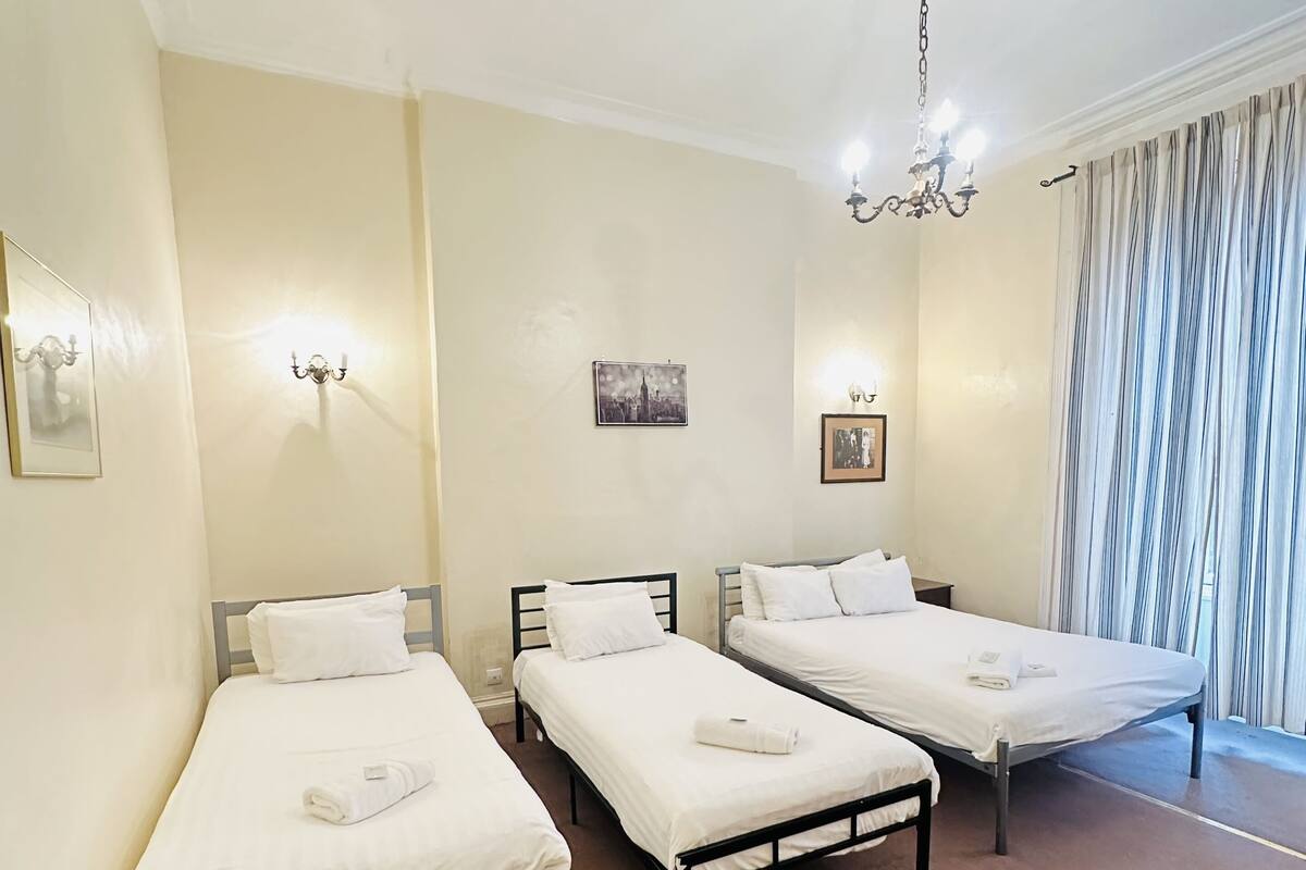 A hotel room with three single beds, a chandelier, and a cityscape picture on the wall.