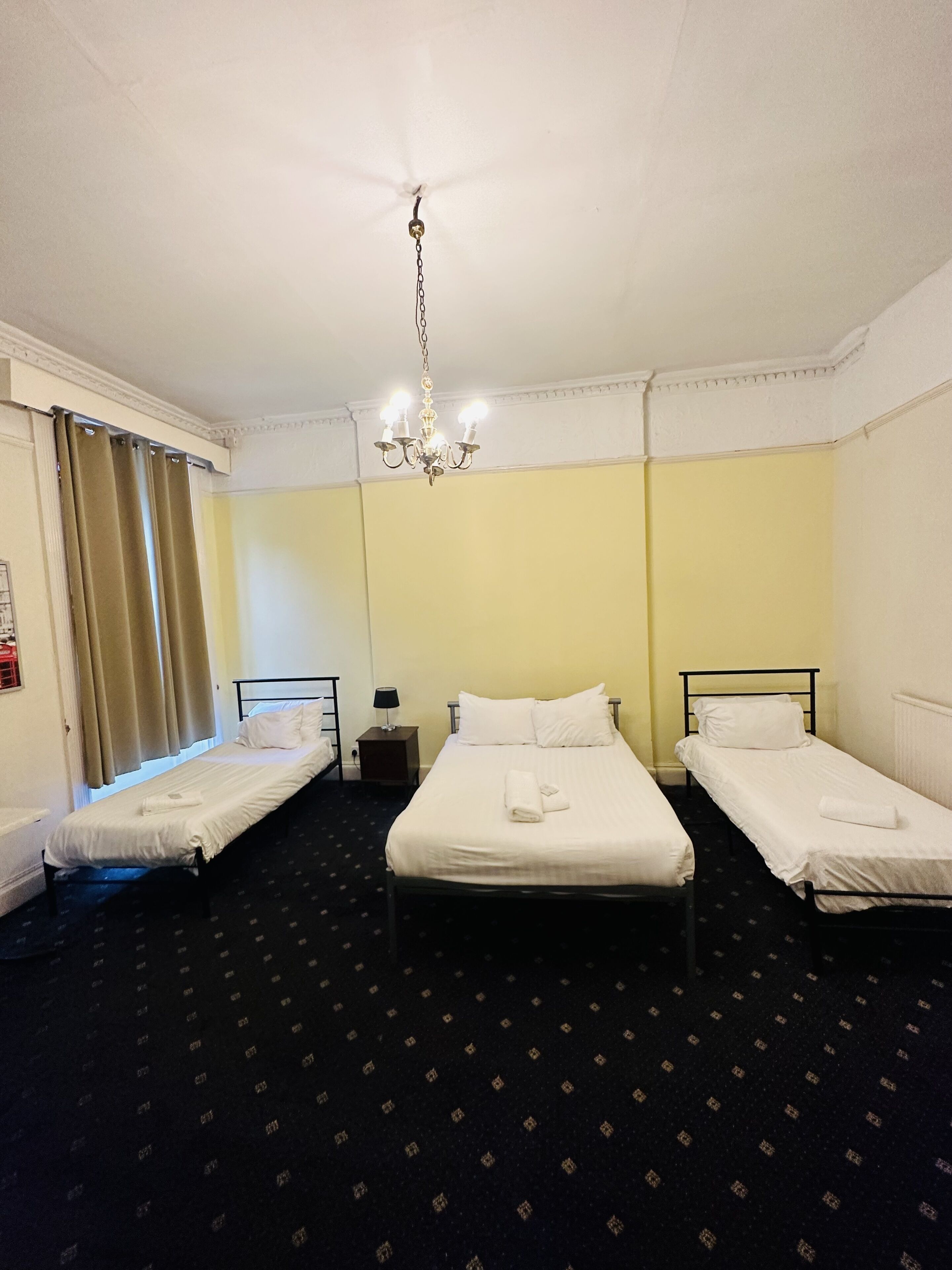 A hotel room with two single beds, a chandelier, a small bedside table with a lamp, and a window with curtains.