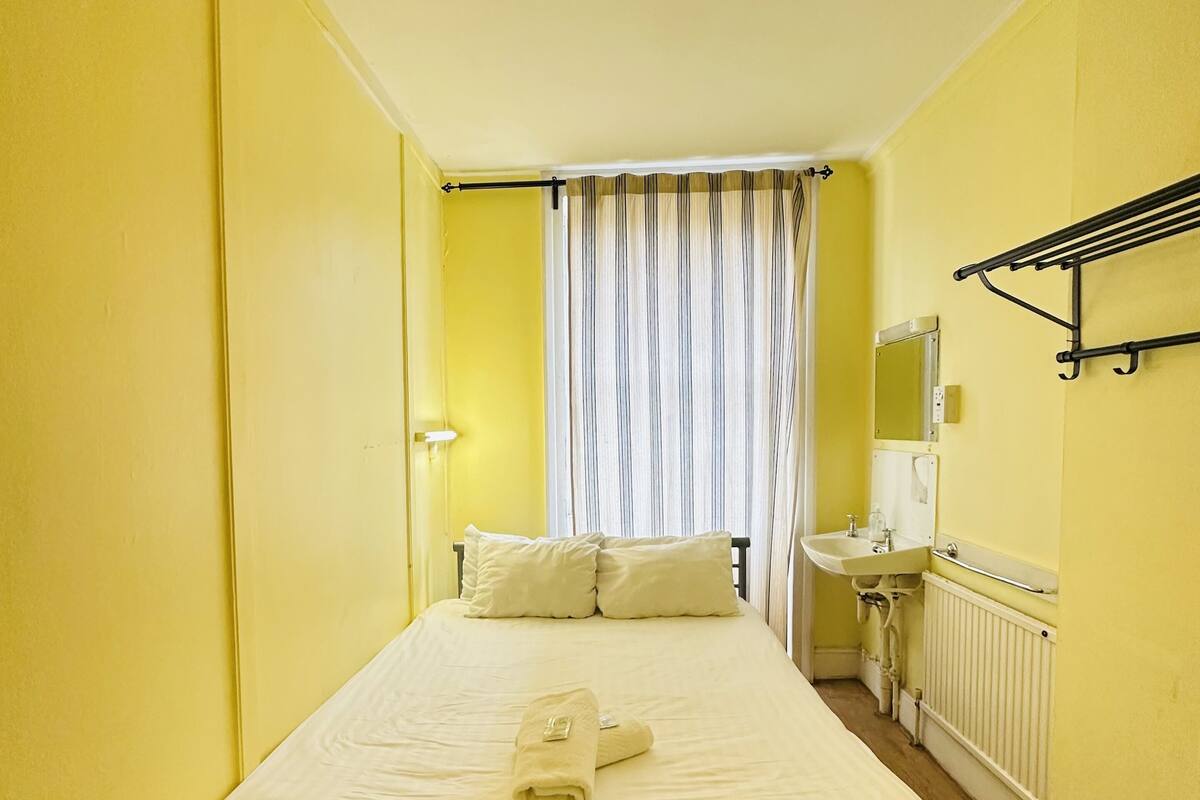 A hotel room with a single bed, a bedside table, a mirror, a radiator, and a window with curtains.