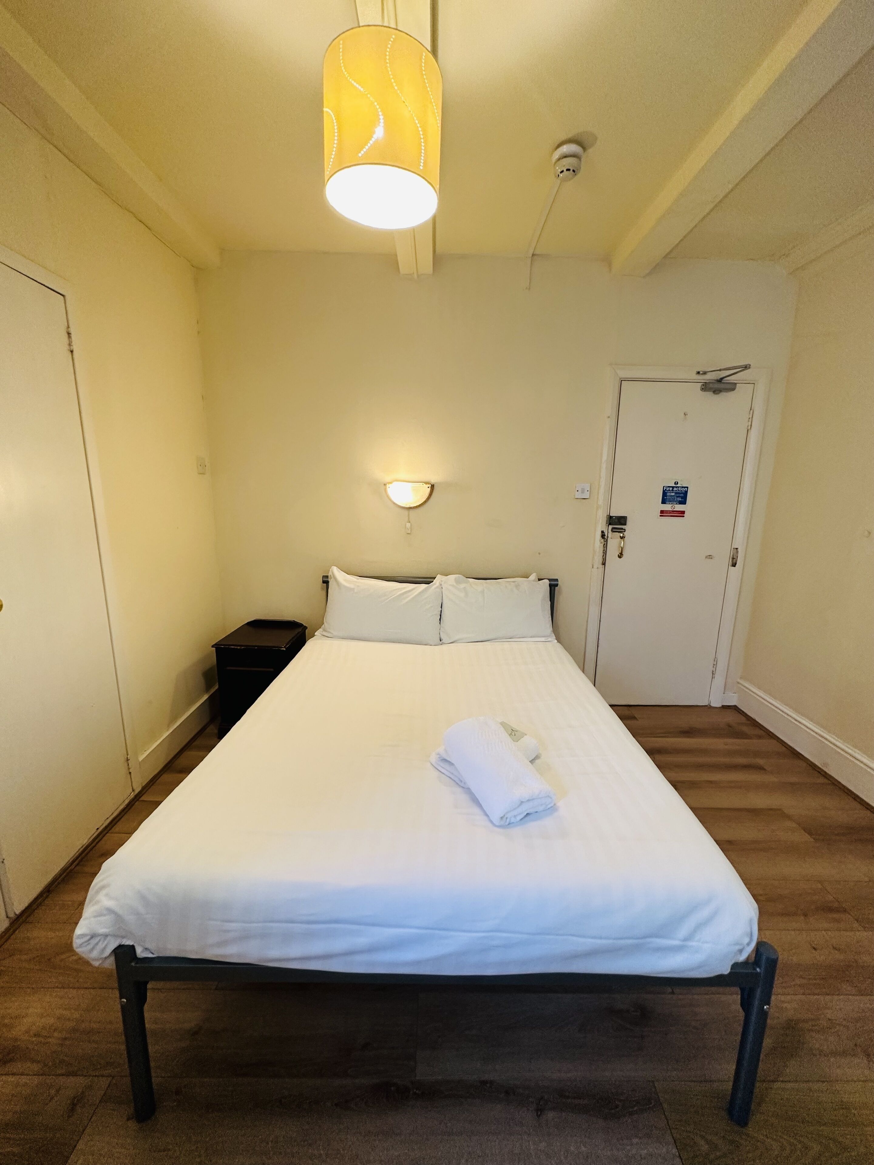 Une chambre d’hôtel comprenant un lit, une table de chevet, une lampe fixée au mur, une porte avec un panneau indiquant les consignes de sécurité incendie et un luminaire au plafond.