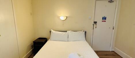 Une chambre d’hôtel comprenant un lit, une table de chevet, une lampe fixée au mur, une porte avec un panneau indiquant les consignes de sécurité incendie et un luminaire au plafond.