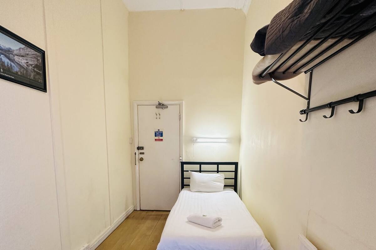 A narrow room with a single bed, a white door, a radiator, and a coat rack.