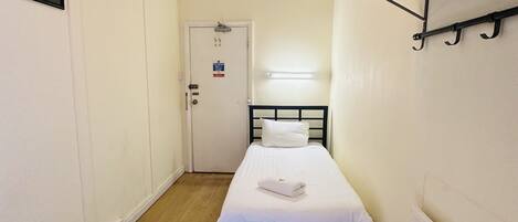A narrow room with a single bed, a white door, a radiator, and a coat rack.