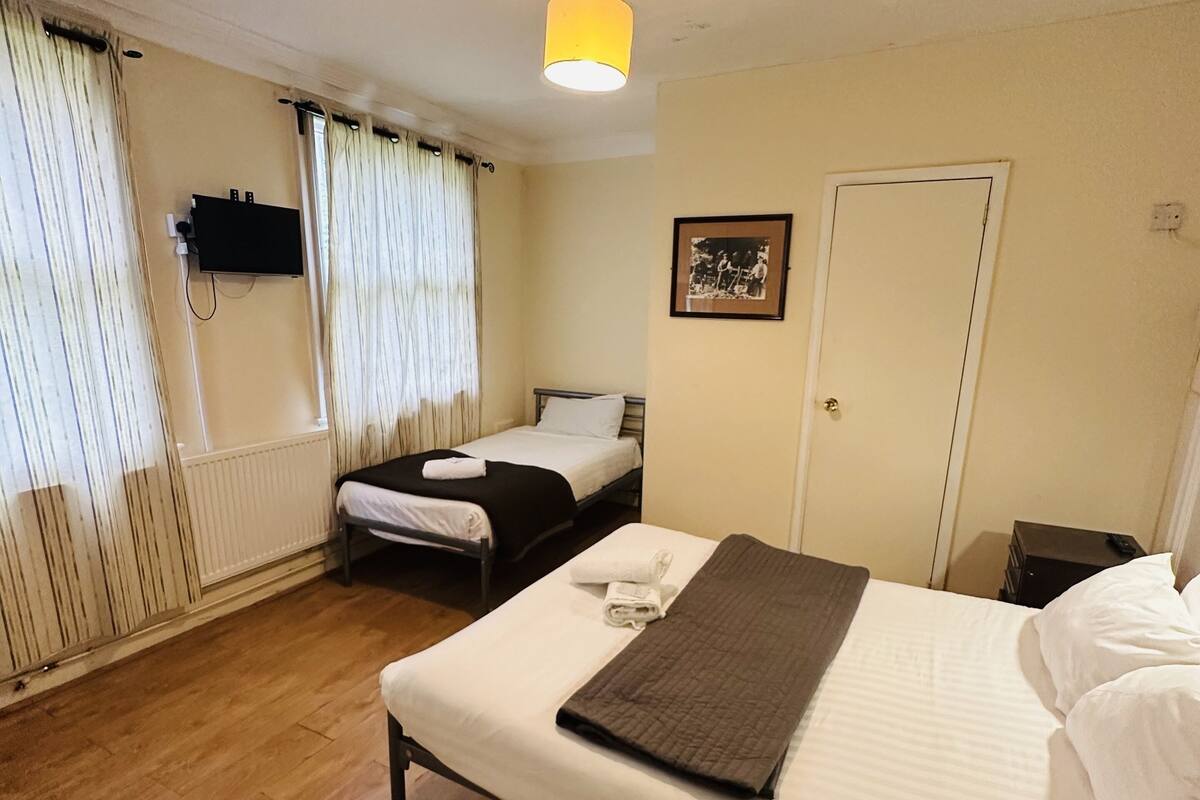 A hotel room with a large bed, a smaller bed, a flat-screen TV mounted on the wall, a bedside table, and a window with curtains.