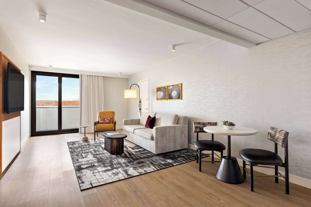 A modern living room with a sofa, armchairs, a coffee table, and a dining table. There is a large window with a view, a flat-screen TV mounted on the wall, and a floor lamp.