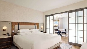 Een moderne hotelkamer met een groot bed, nachtkastjes en een uitzicht door een schuifdeur op een eetruimte.