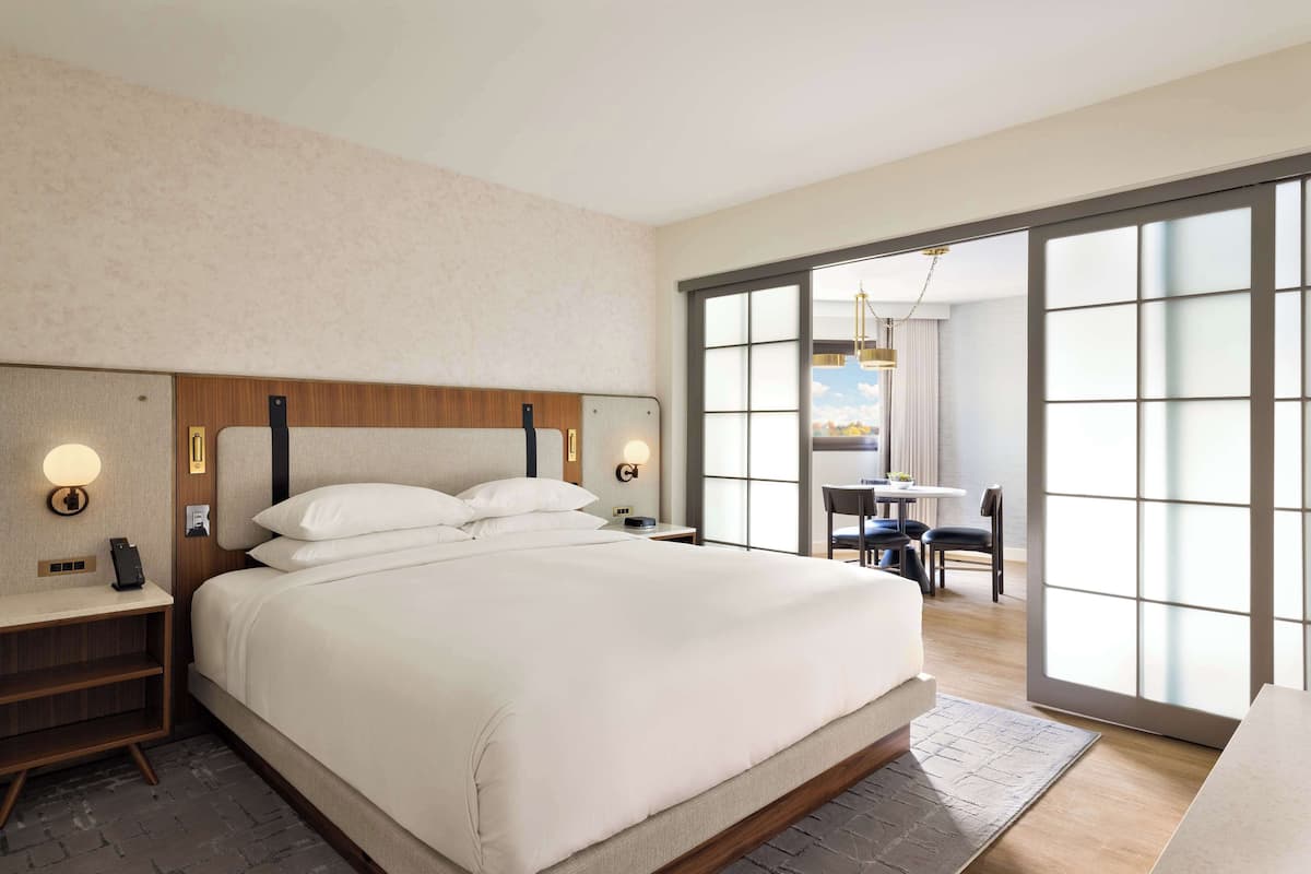 A modern hotel room with a large bed, bedside tables, and a view through a sliding glass door to a dining area.
